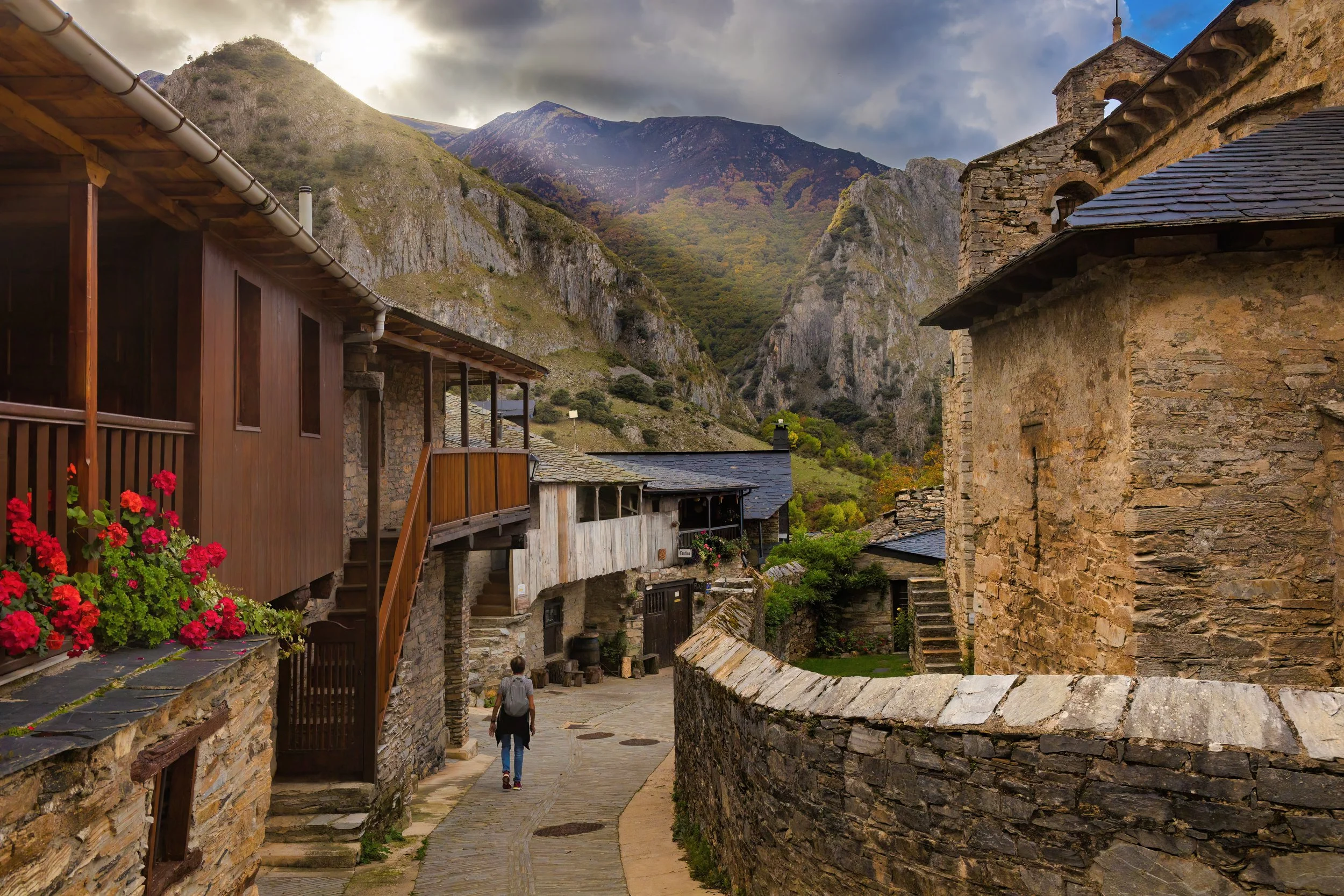 Vista de un pequeño pueblo en un valle rodeado de montañas, con casas de piedra y madera, una calle empedrada y un cielo nublado.