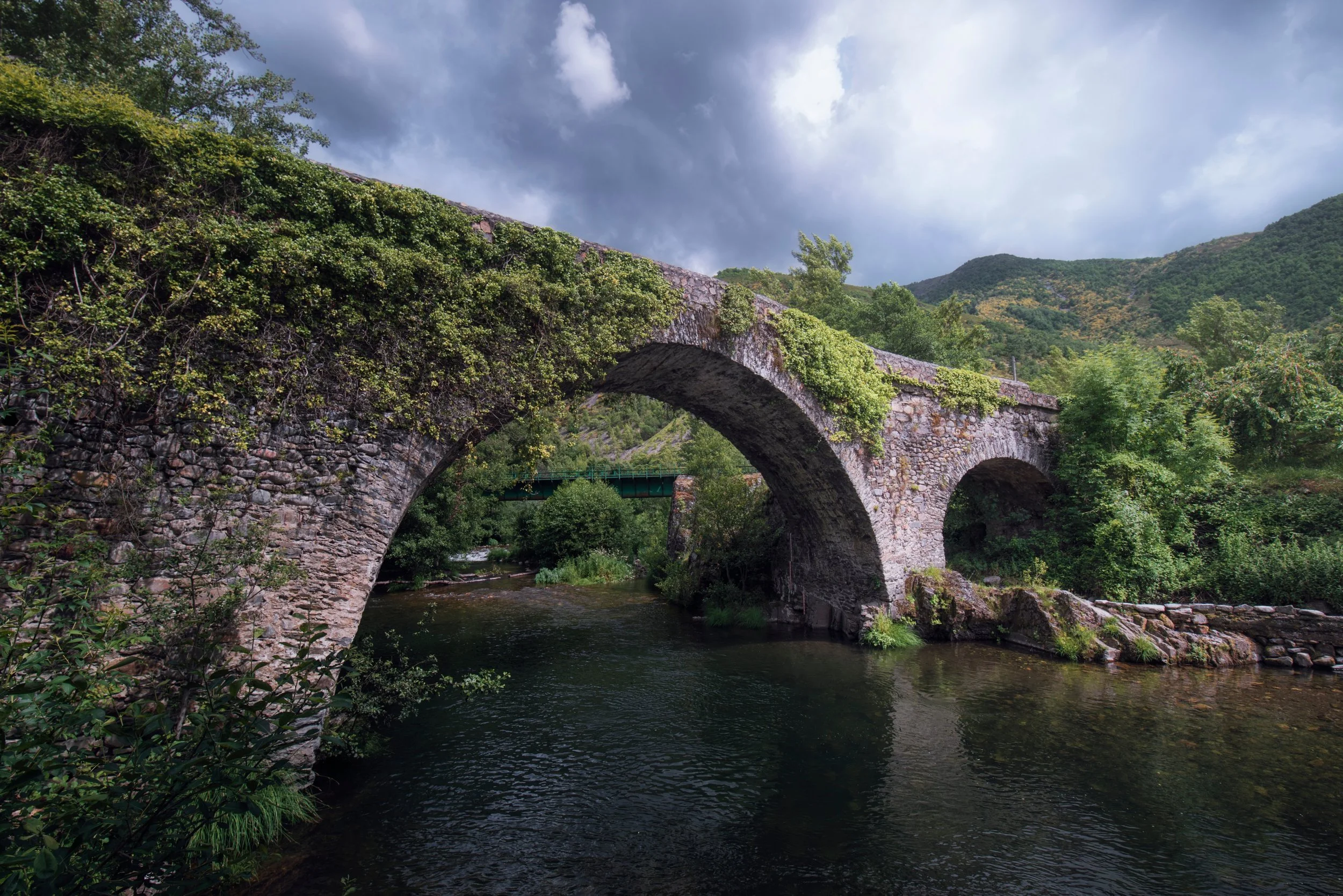Puente de piedra cubierto de vegetación sobre río en un paisaje montañoso nublado en el fondo.