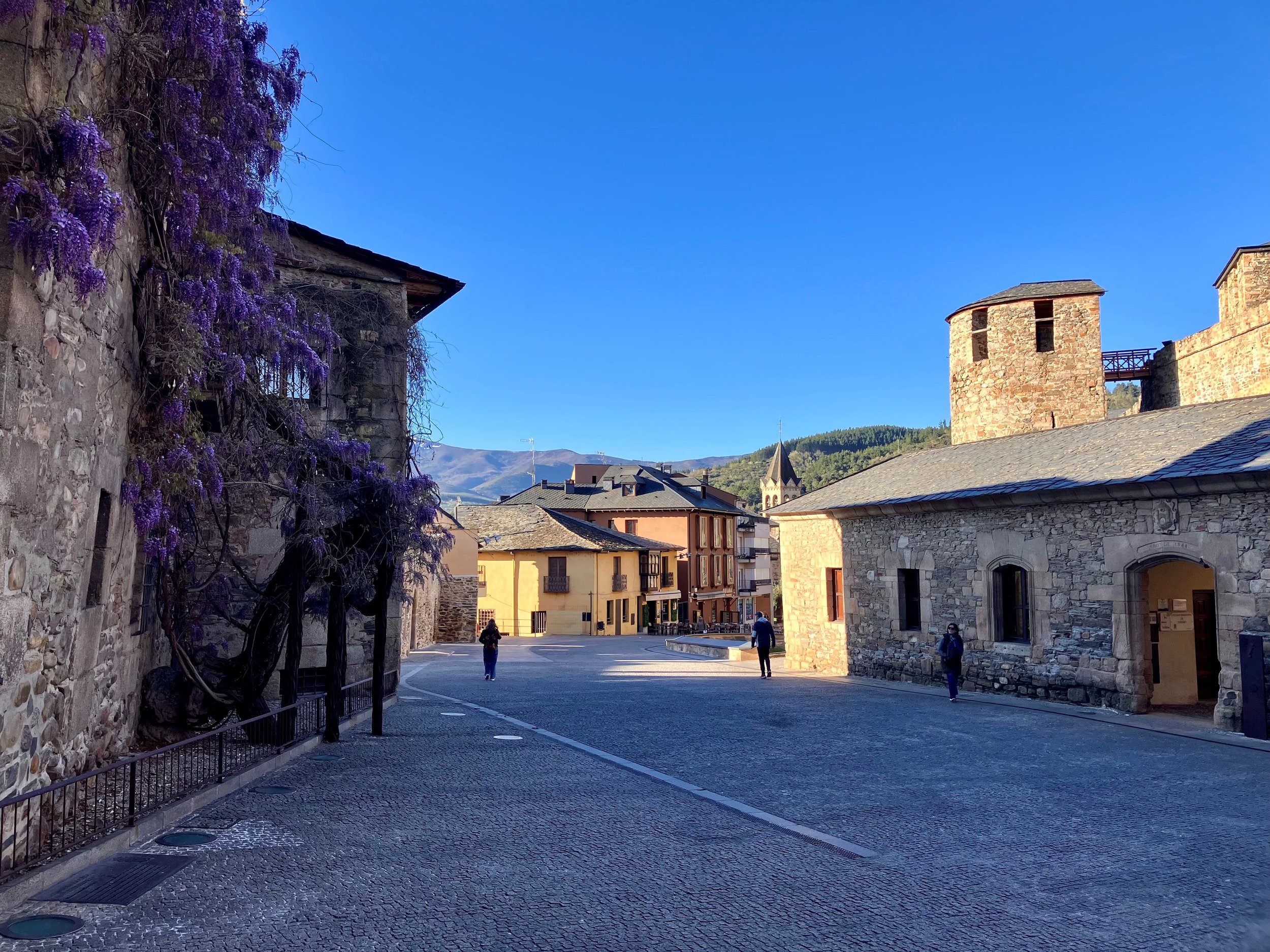Calles de un pueblo con edificios de piedra y techos de teja en un día despejado, montañas al fondo, un árbol con flores moradas, varias personas caminando.