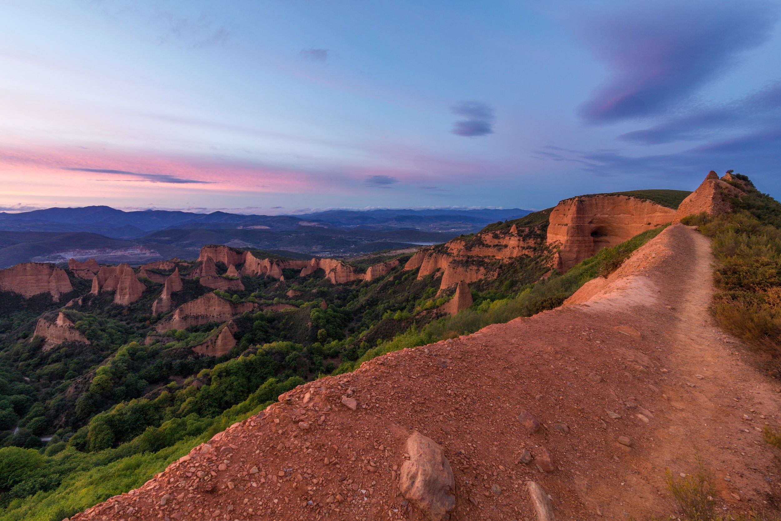 Paisaje con formaciones rocosas, colinas verdes y un cielo con tonos rosados y azules en el atardecer.