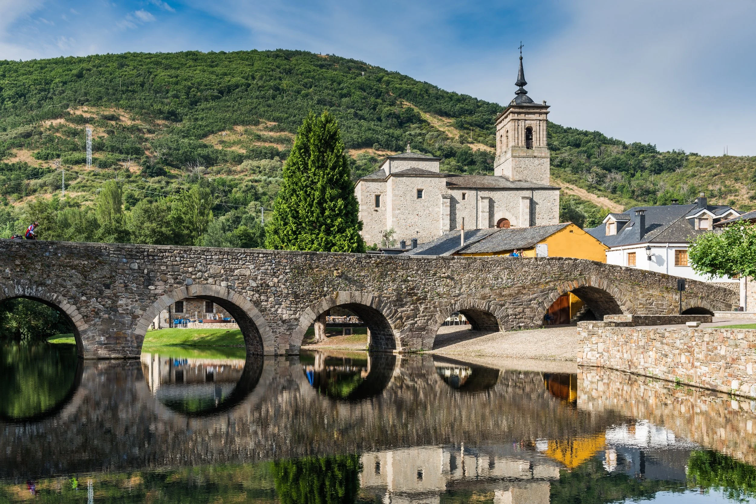 Puente de piedra sobre un río refleja el castillo y las casas de una ciudad medieval en un entorno montañoso con vegetación verde y cielo azul con nubes
