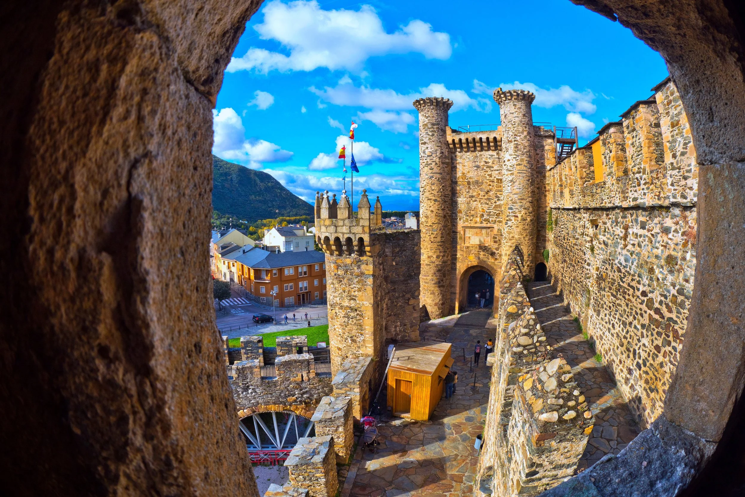 Vista de una antigua fortaleza de piedra, vista desde una ventana ovalada, con un cielo azul y nubes, y un pueblo y montañas en el fondo.