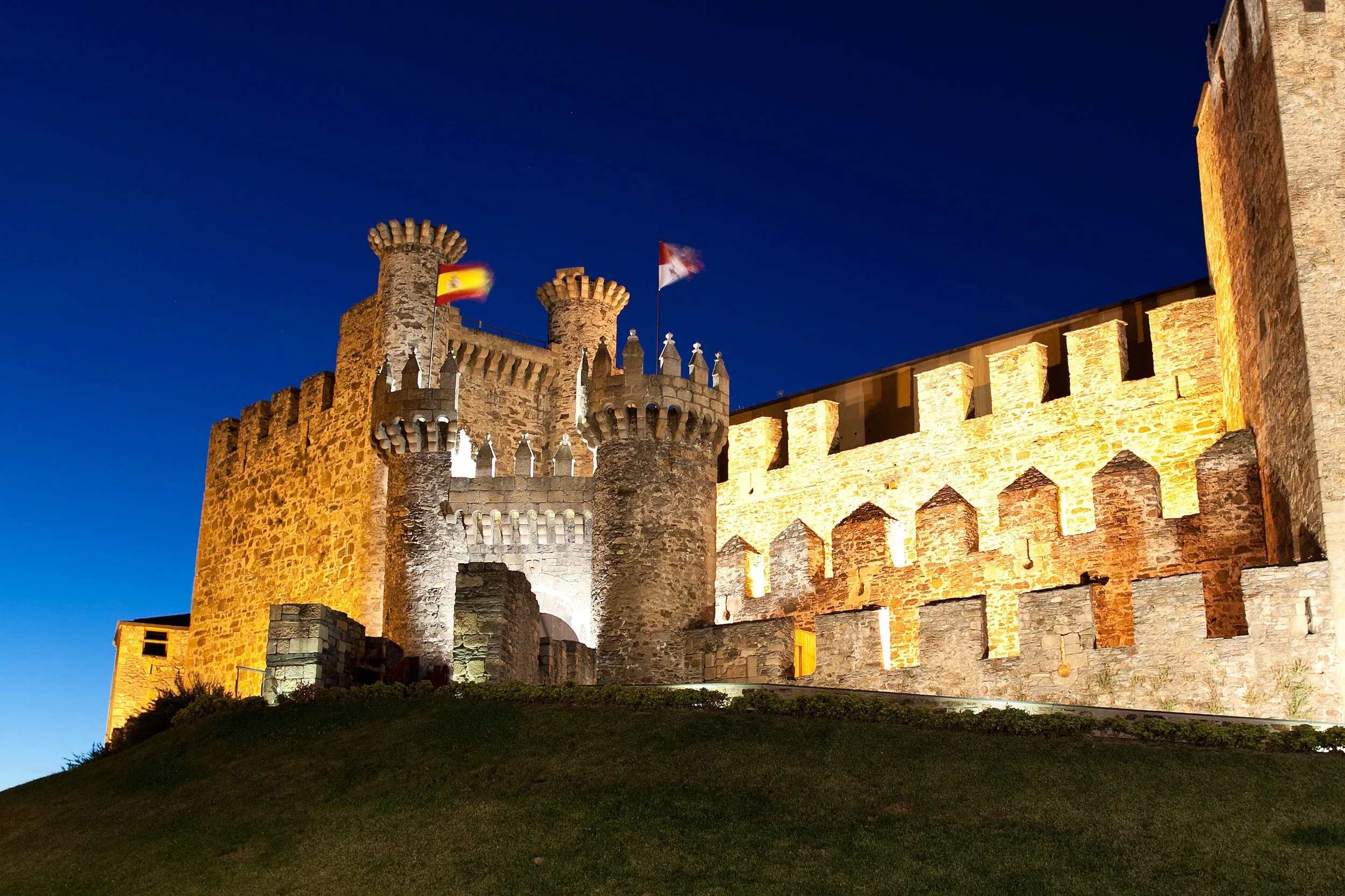 Castillo medieval de piedra iluminado por la noche, con banderas en lo alto de las torres y un cielo oscuro de fondo.