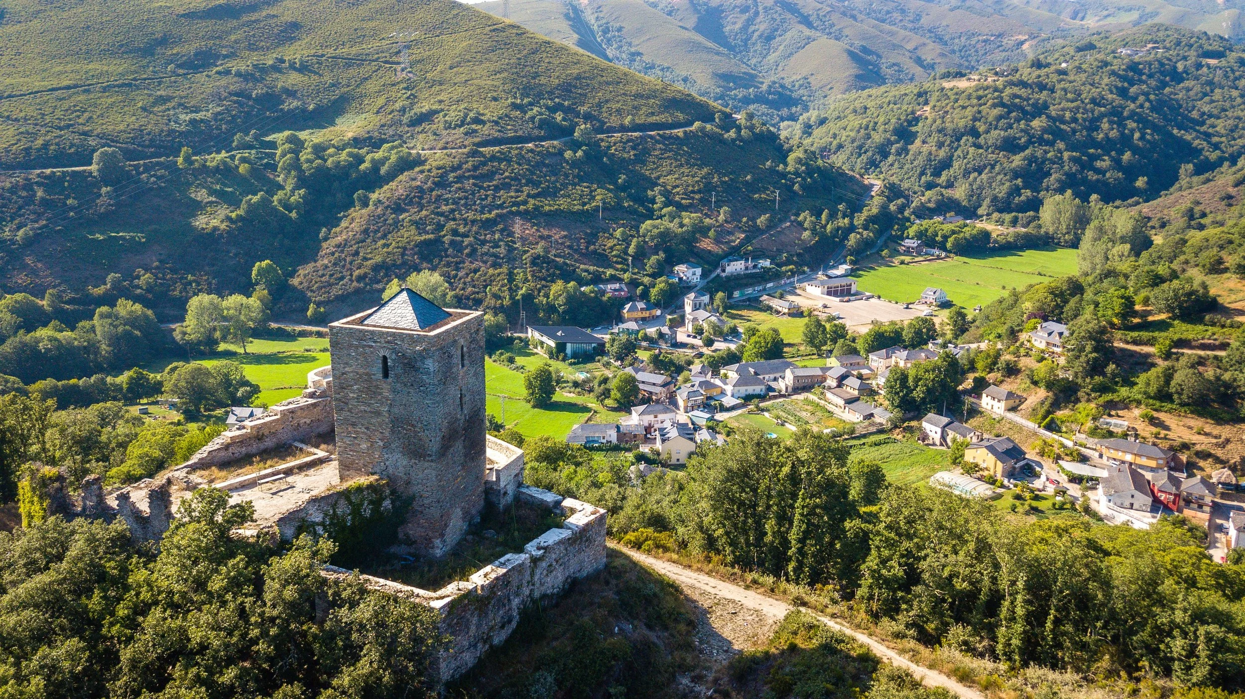 Vista panorámica de una aldea rodeada de montañas verdes, con una torre antigua de piedra en primer plano.