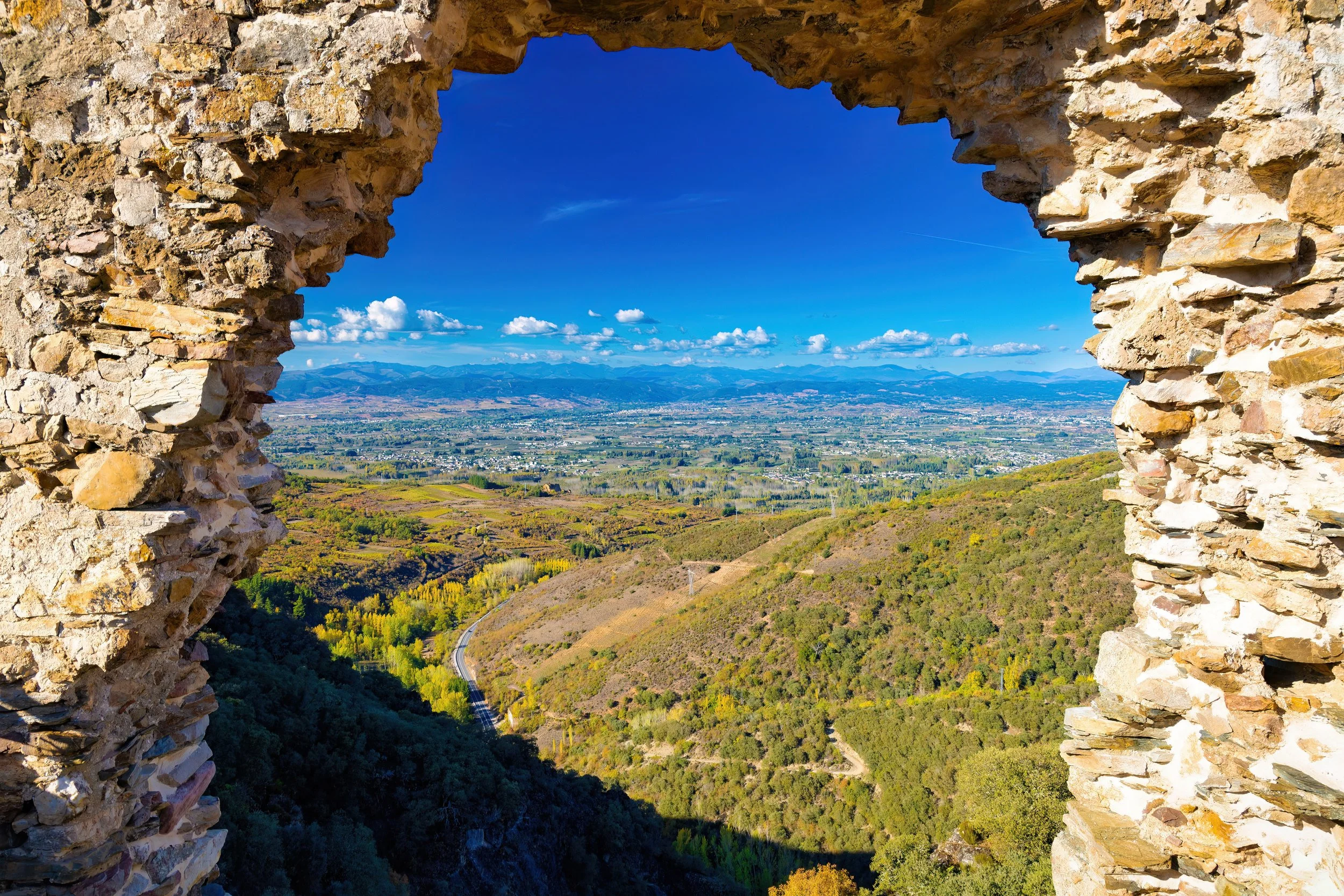 Vista panorámica de un valle con campos y montañas al fondo, vista a través de una ventana en una estructura de piedras.
