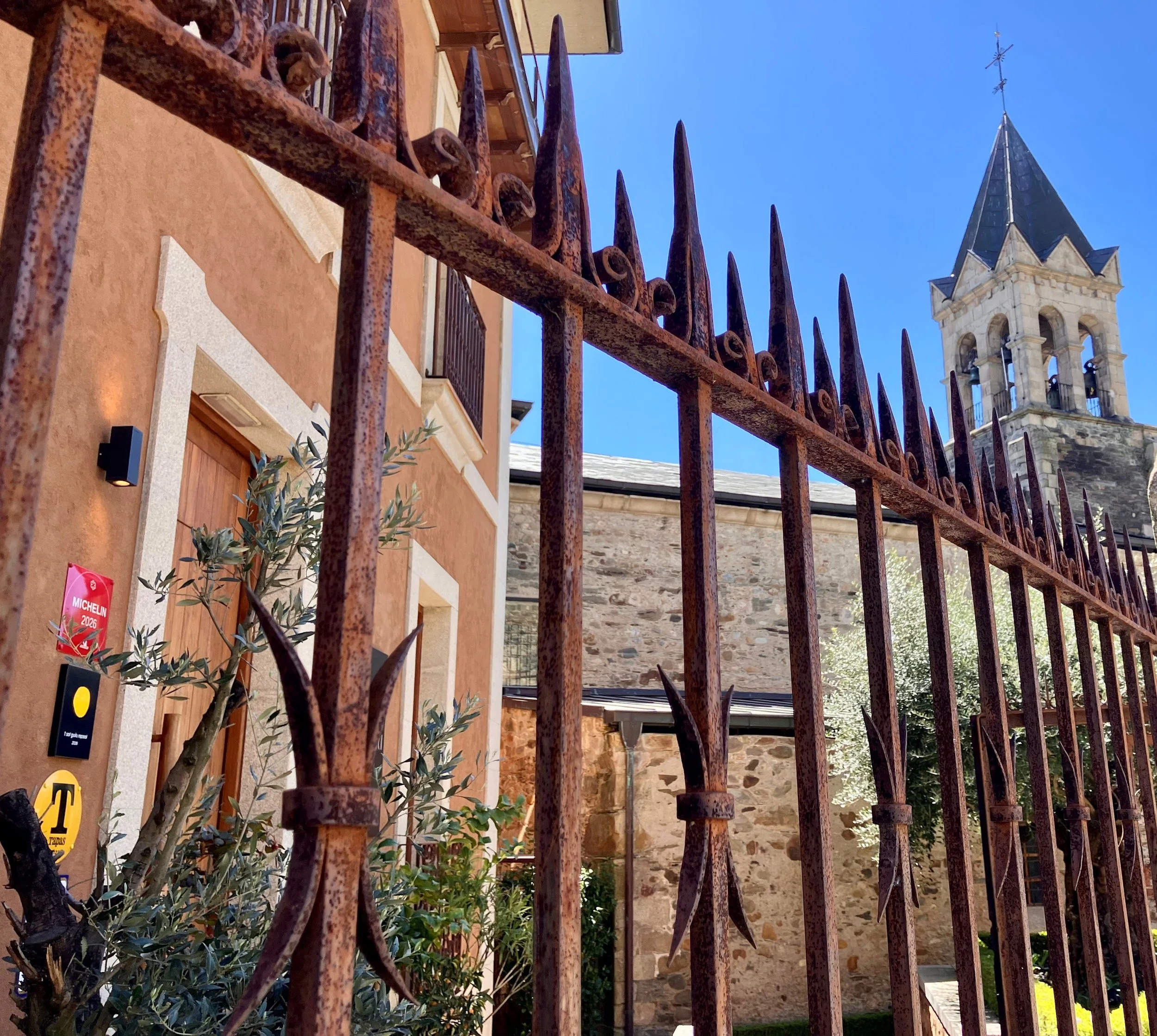 Una verja de hierro con pinchos oxidados, una iglesia con campanario y una cruz en la parte superior, y un edificio de color ocre con ventanas y plantas decorativas en un día soleado con cielo azul.