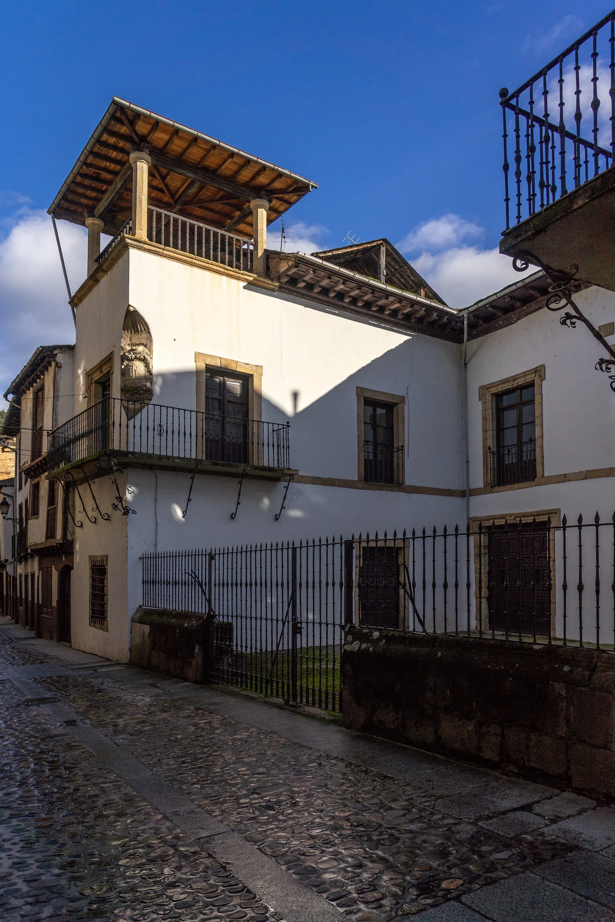 Edificio clásico con paredes blancas y ventanas de madera, rejas de hierro y un balcón en la fachada. El cielo azul y algunas nubes aparecen en el fondo, y la calle de adoquines en primer plano refleja la luz del atardecer.