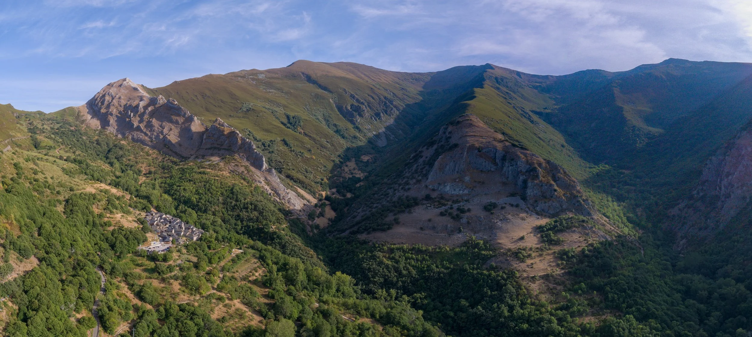 Paisaje de montañas verdes con algunos picos rocosos y una pequeña aldea en la parte inferior izquierda, bajo un cielo despejado con algunas nubes delgadas.