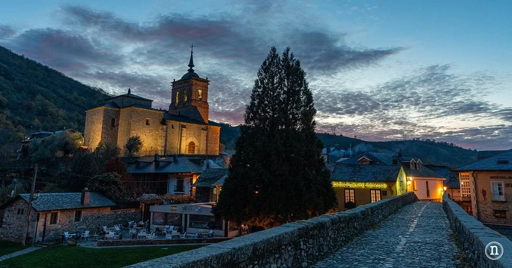 Vista de Molinaseca con una iglesia antigua en una colina, árboles y casas, al anochecer.