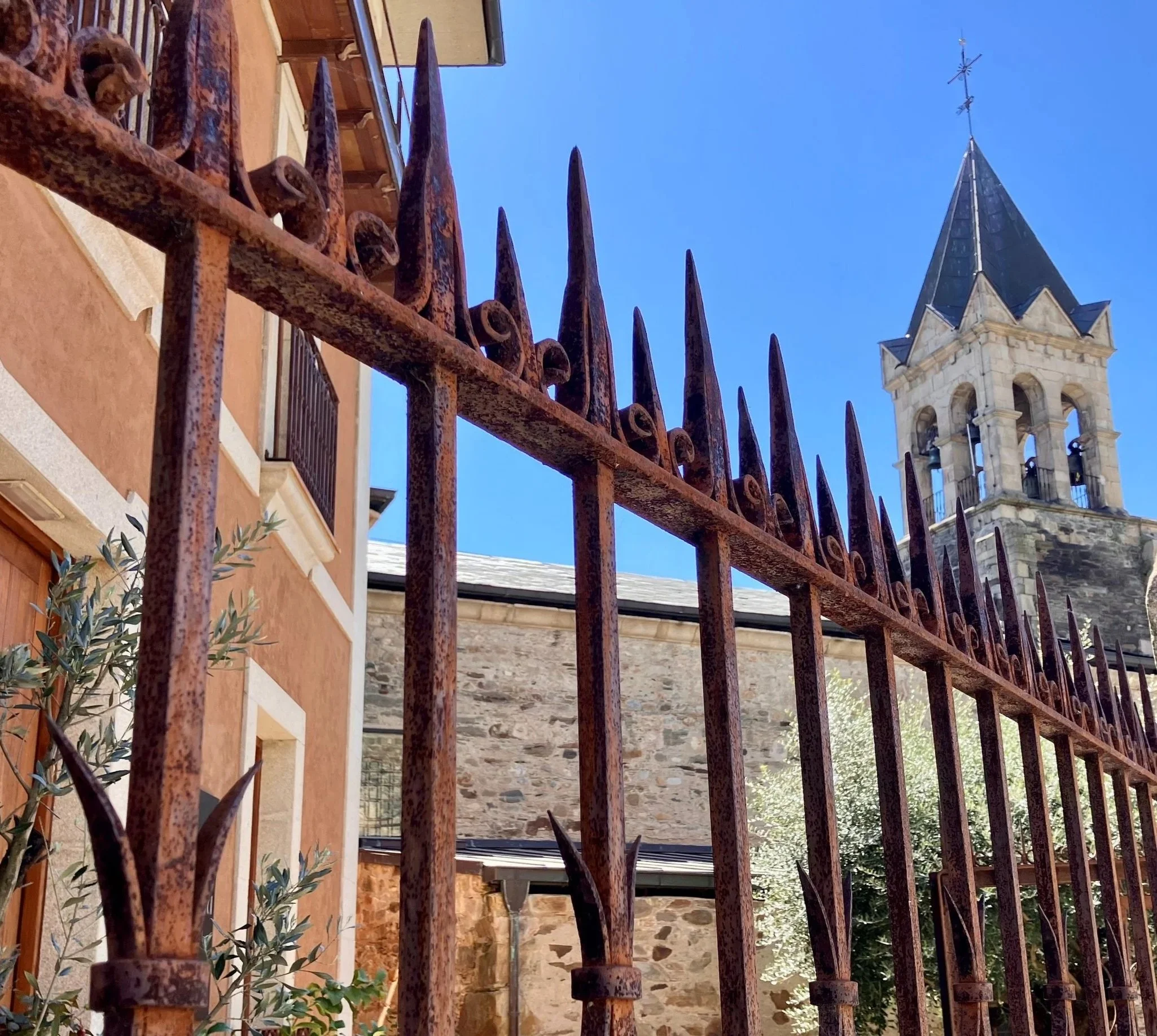 Barandilla de hierro forjado oxidada en primer plano, con iglesia o torre antigua de piedra y tejado puntiagudo al fondo, todo bajo cielo azul claro.