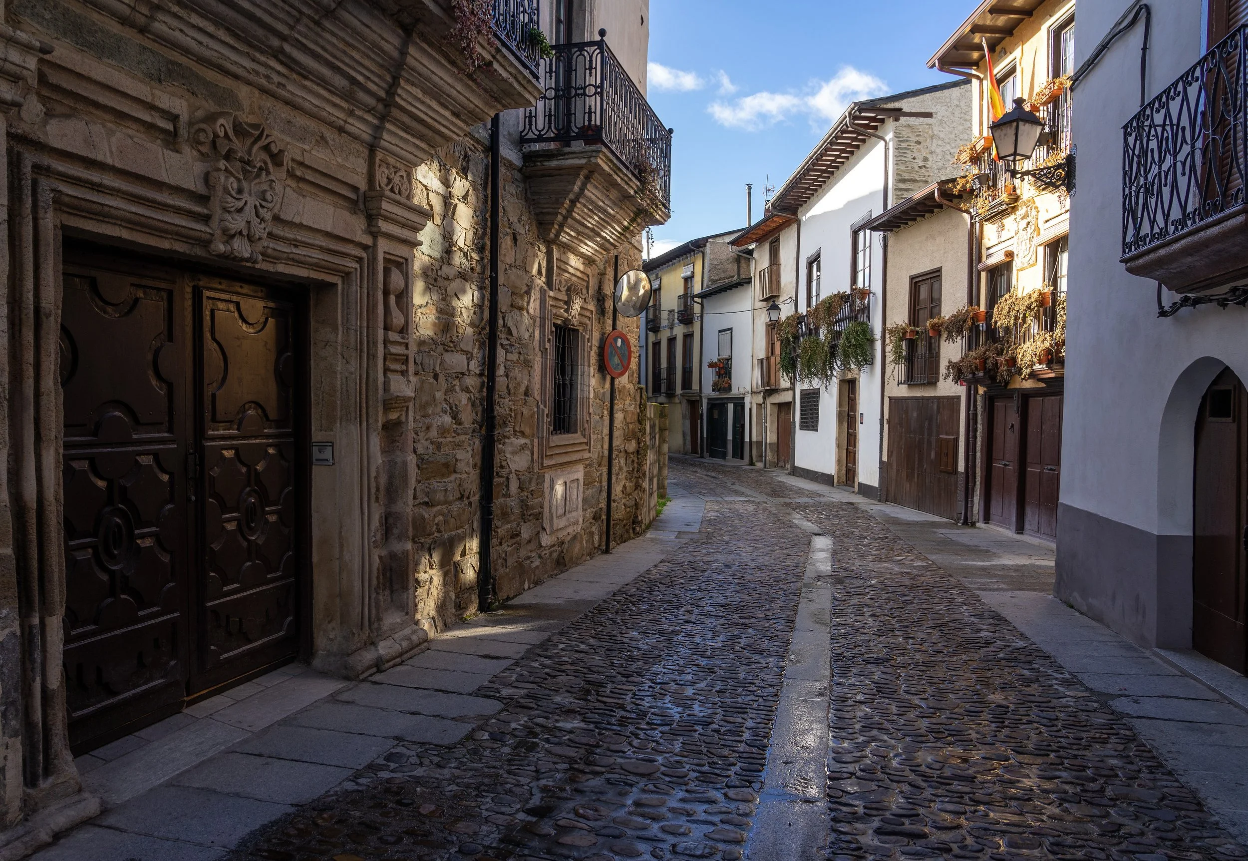 Calles empedradas de una ciudad antigua con edificios de paredes de piedra y pisos de piedra, balcones con rejas de hierro y ventanas con macetas, bajo un cielo despejado.