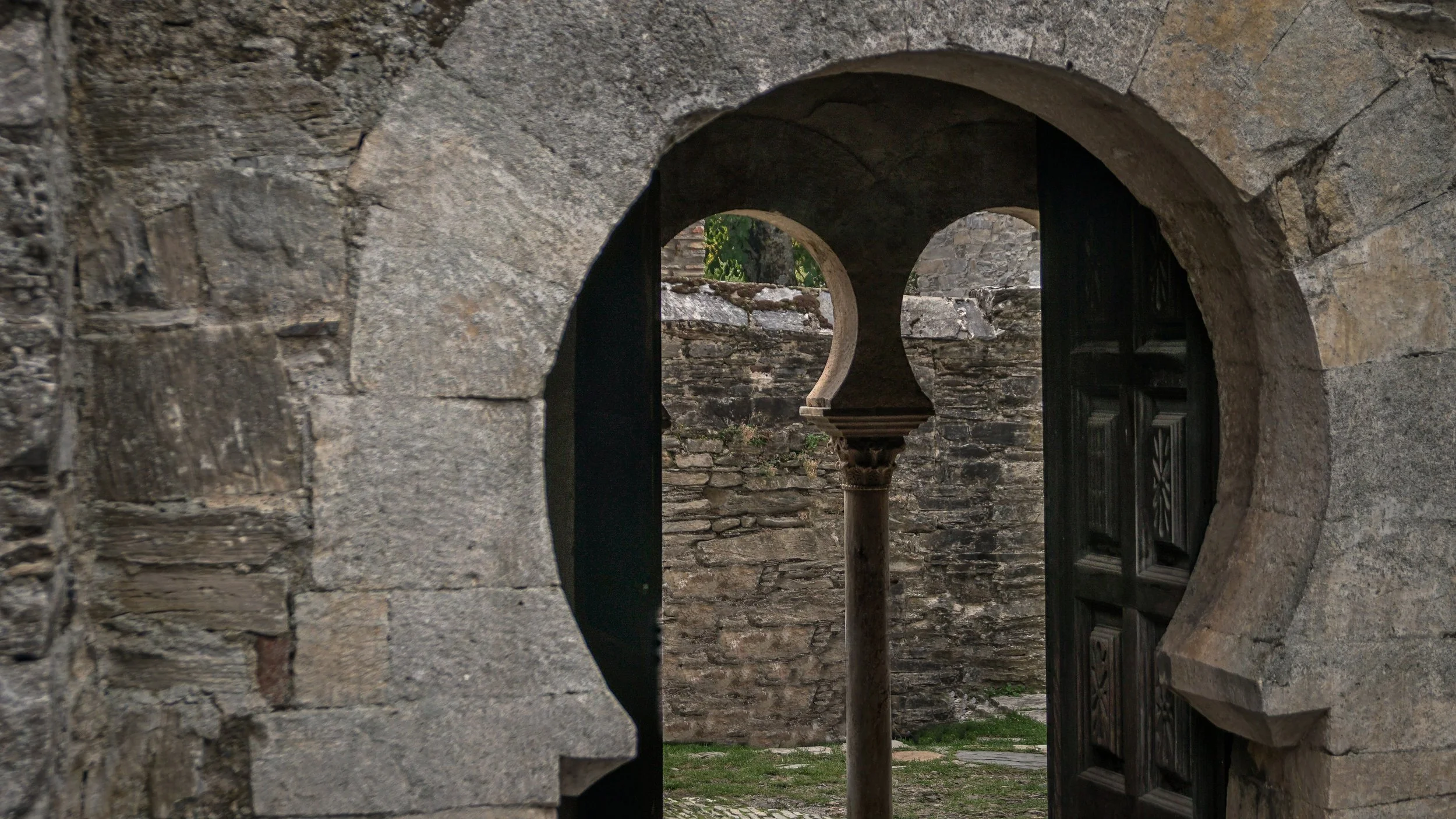 Vista a través de una abertura en una pared de piedra, mostrando un patio con columnas y paredes antiguas, típicas de una construcción histórica o antigua.