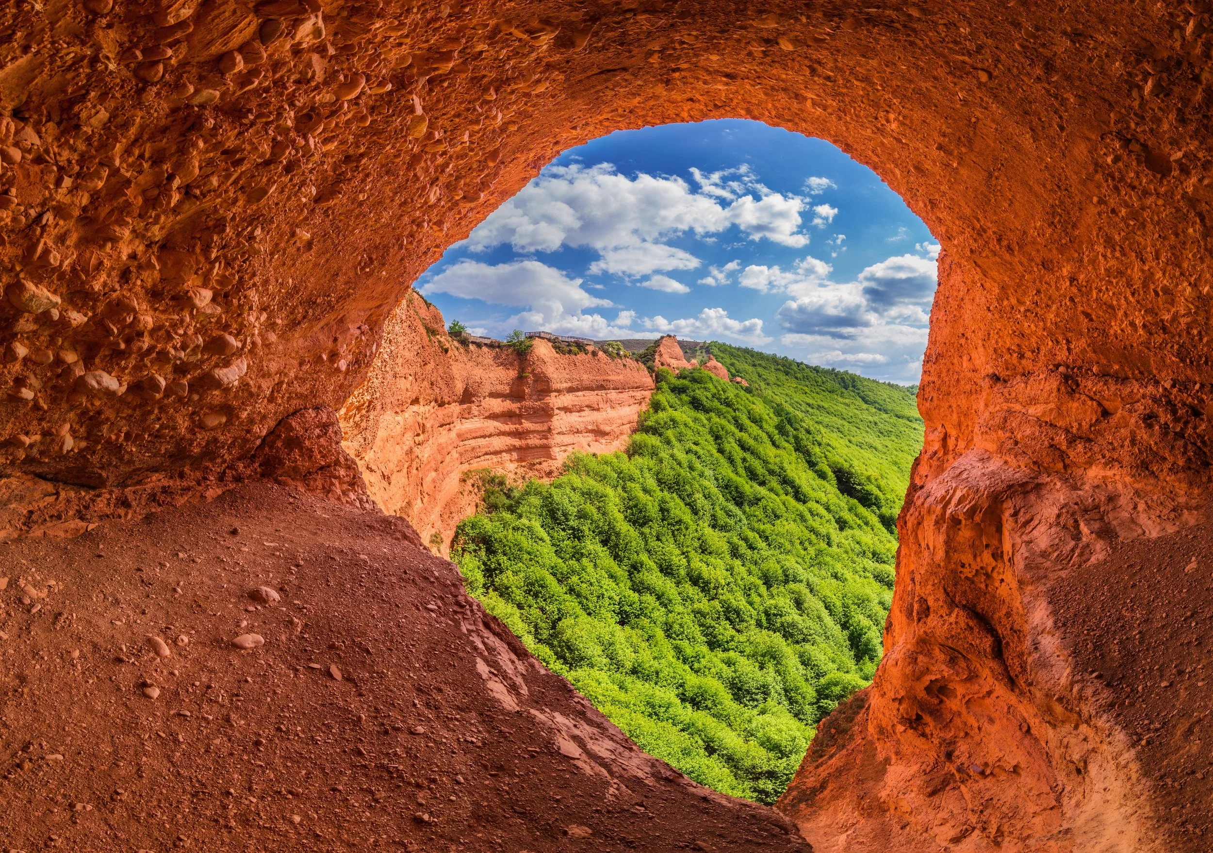 Vista desde dentro de una cueva roja mirando hacia un cielo azul con nubes y un paisaje verde con colinas.