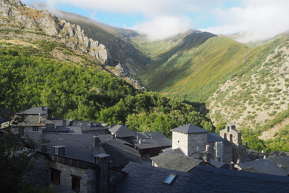 Vista de un pueblo montañoso con techos de teja gris, rodeado de vegetación y montañas verdes con nubes en el cielo.