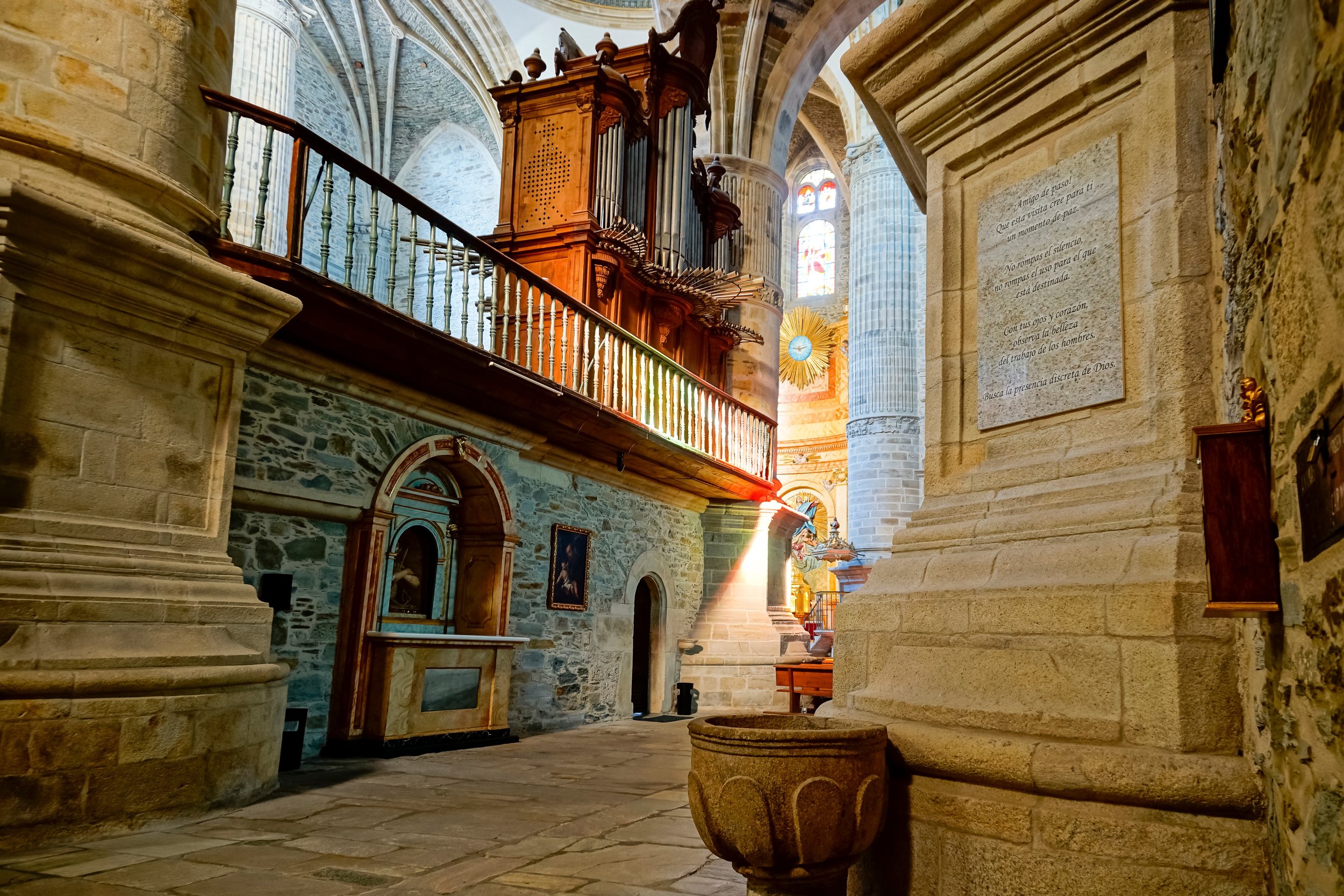 Interior de una iglesia con columnas de piedra, un órgano de madera en un segundo nivel, vitrales y un altar con un sol dorado con un reloj en el centro, en luz natural que entra por las ventanas.