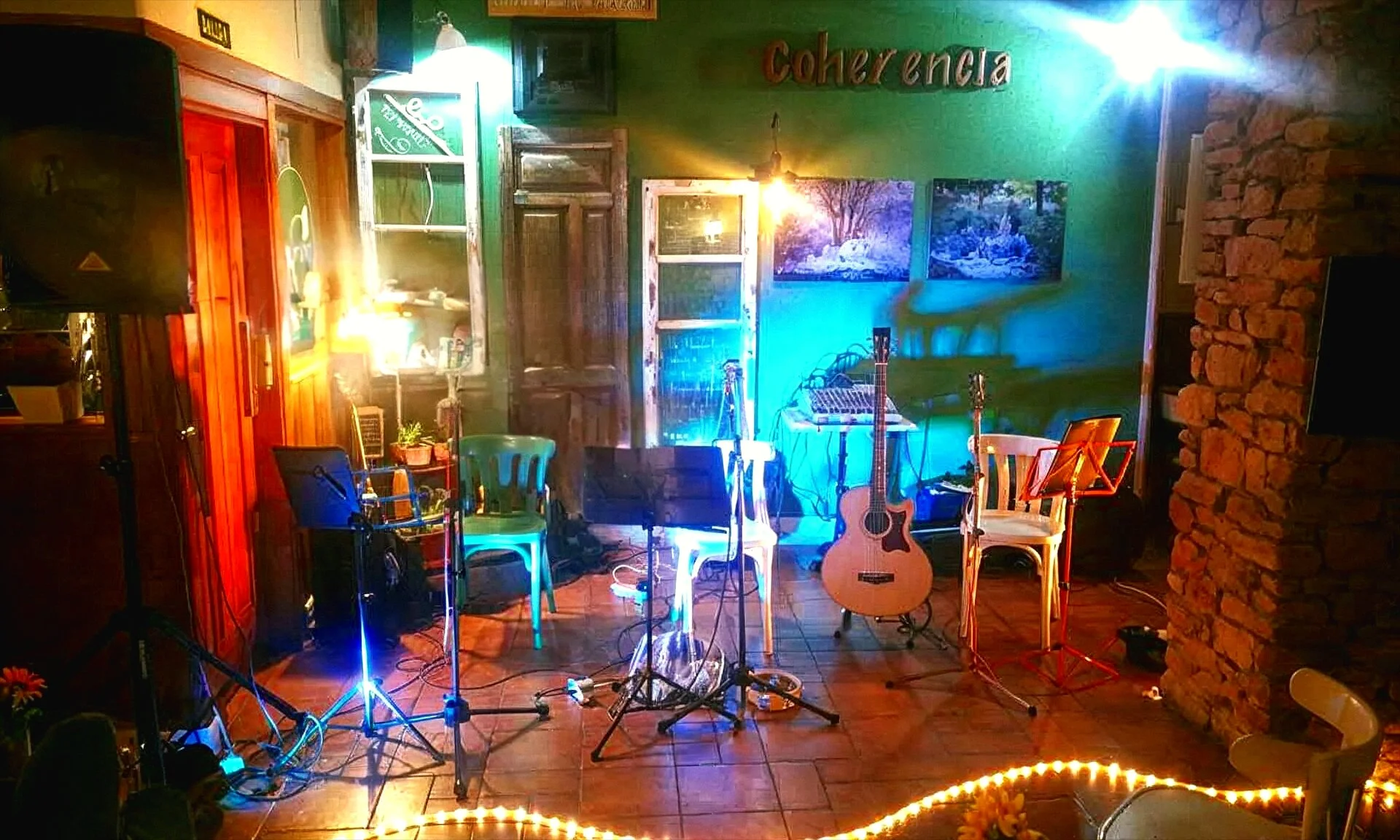 Un escenario musical en un establecimiento llamado Coherencia, con guitarras, sillas, luces coloridas y decoraciones acusticas en las paredes.