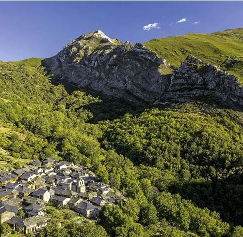 Vista de un pueblo pequeño con casas y techos grises, rodeado de árboles verdes y montañas al fondo bajo un cielo azul con algunas nubes.