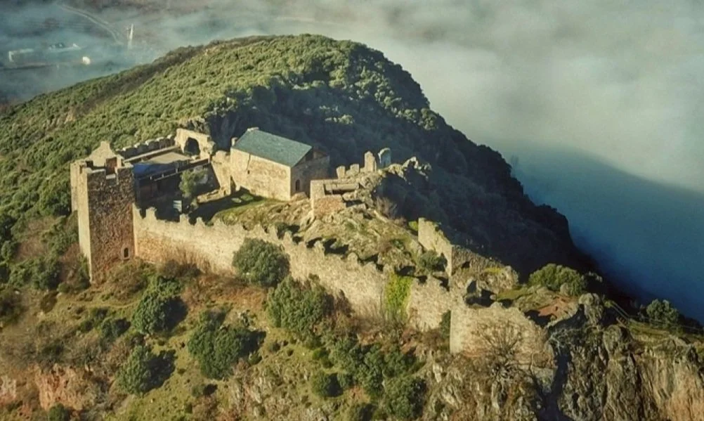 Castillo de Cornatel en una colina cubierta de vegetación, rodeado de murallas y con una estructura de techo azul, en un paisaje montañoso con nubes y niebla.