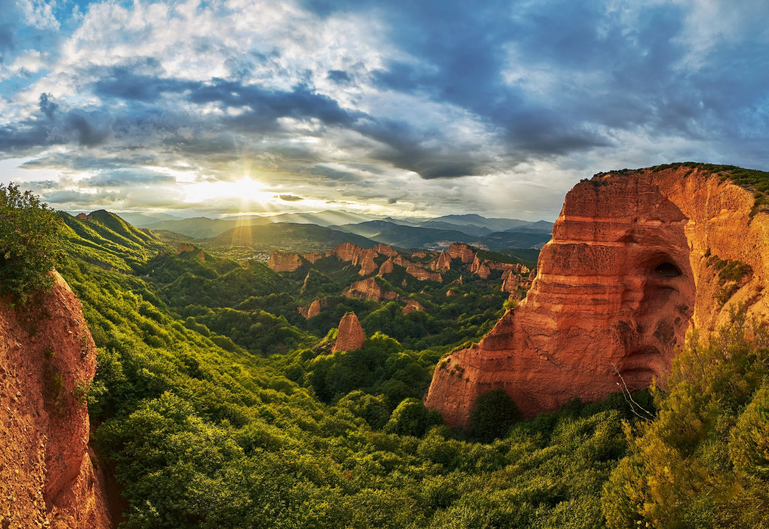 Paisaje de montañas con vegetación y formaciones rocosas rojas, cielo con nubes y sol naciente o poniente.