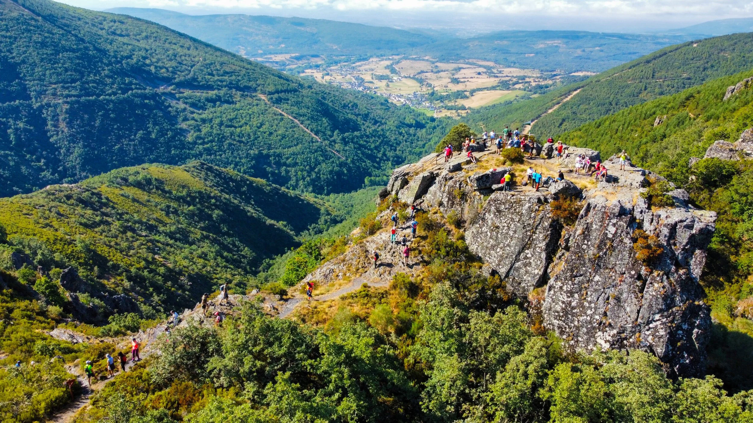 Varias personas haciendo senderismo en una cresta de roca en medio de un valle montañoso cubierto de árboles. Al fondo se observa un paisaje rural con campos y pequeñas localidades.