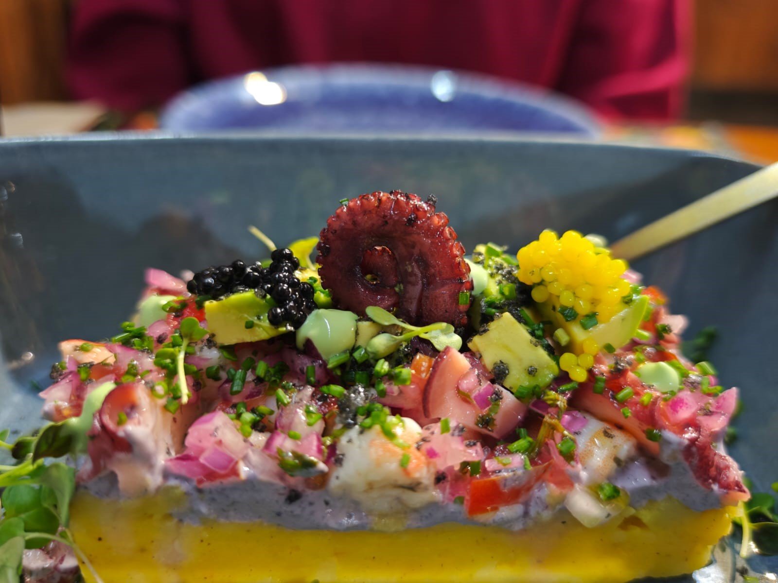 Plato de comida con mariscos, aguacate, cebolla, y varias salsas, adornado con semillas de chía y una rodaja de remolacha en la parte superior