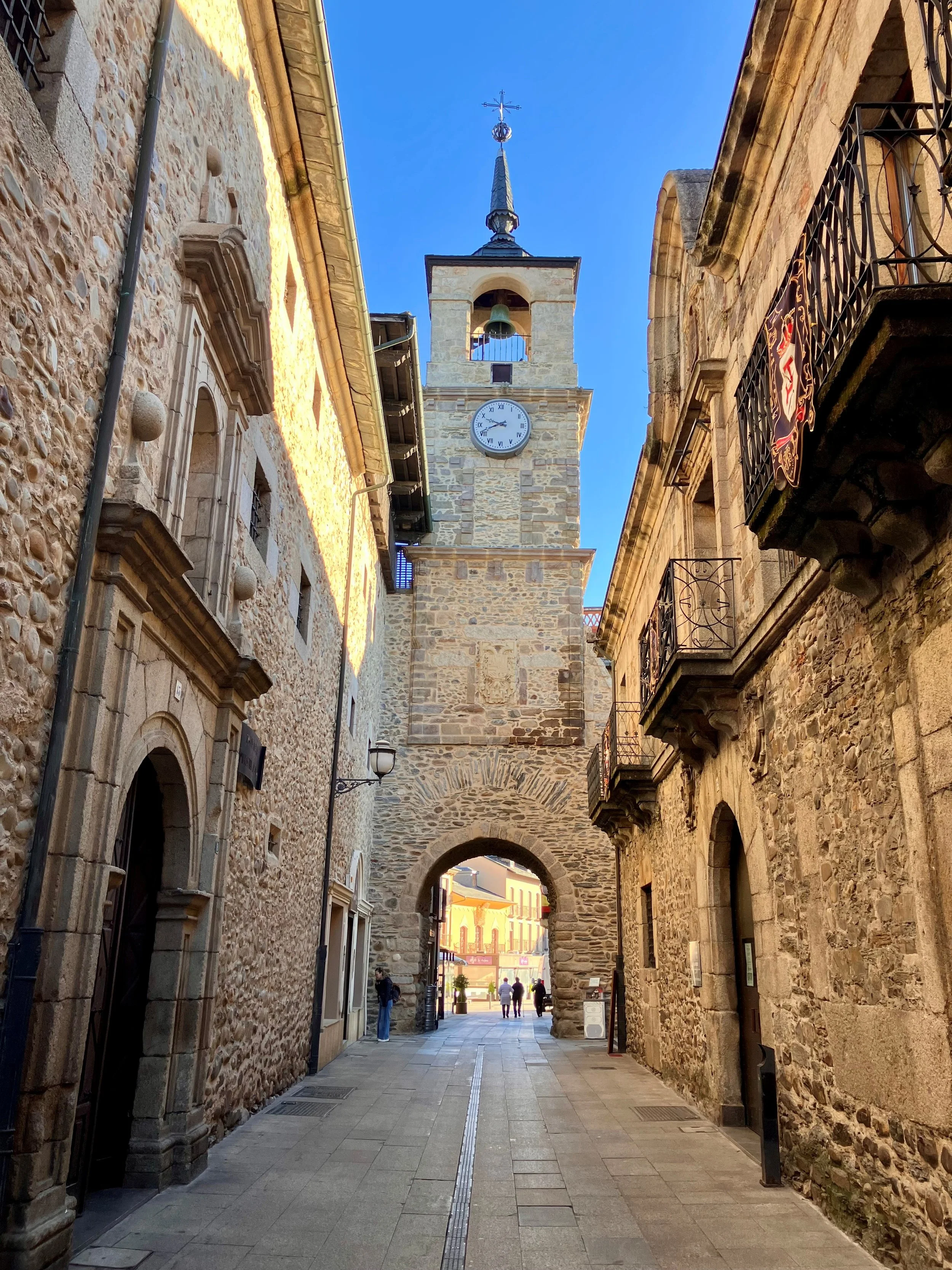 Calles de piedra con un arco en el fondo y una torre de reloj con campana, en una tarde soleada con cielo azul.