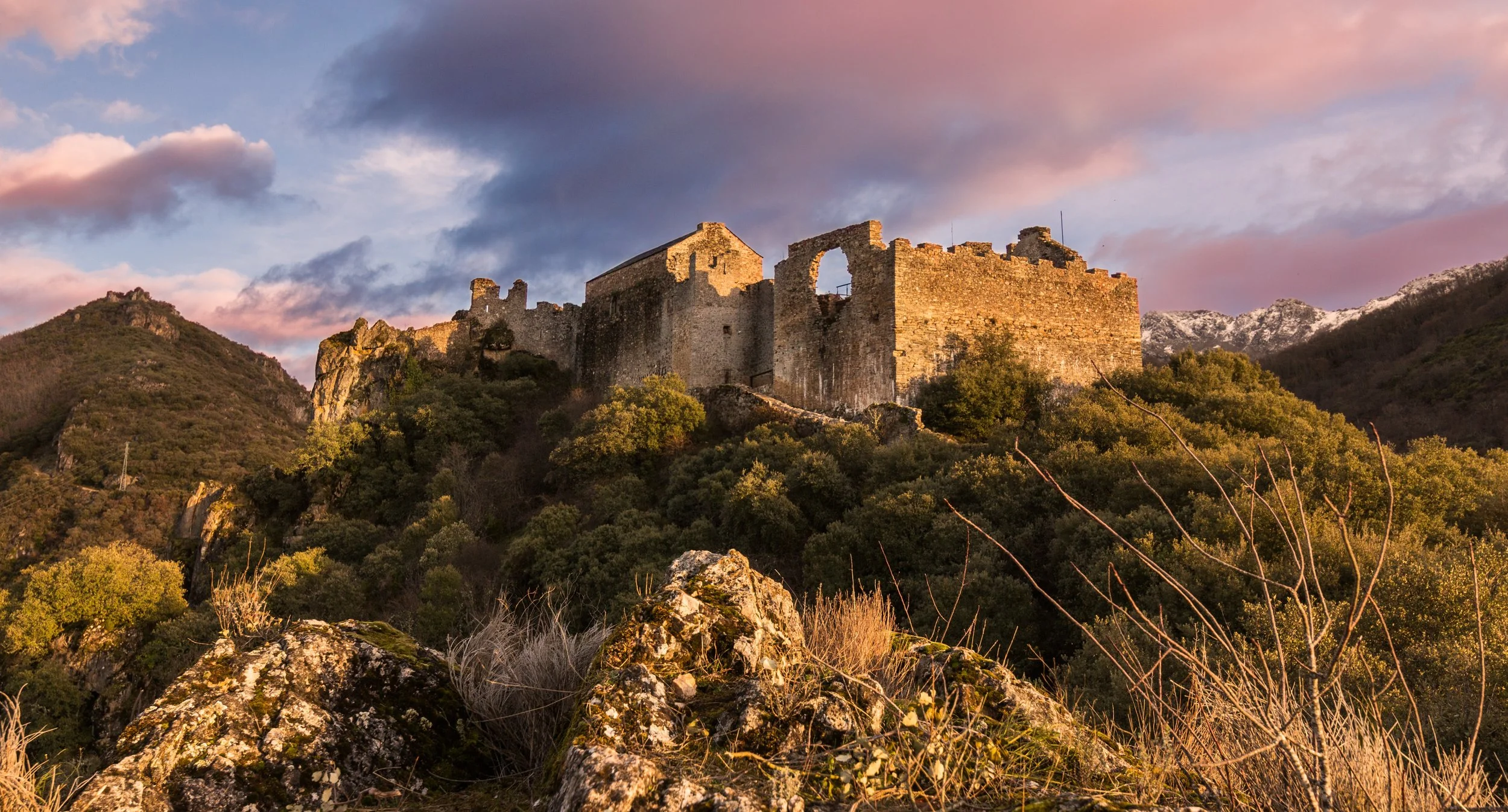 Ruinas de un castillo antiguo en una colina rodeado de naturaleza y montañas con nieve en el fondo, durante el atardecer con cielo colorido.
