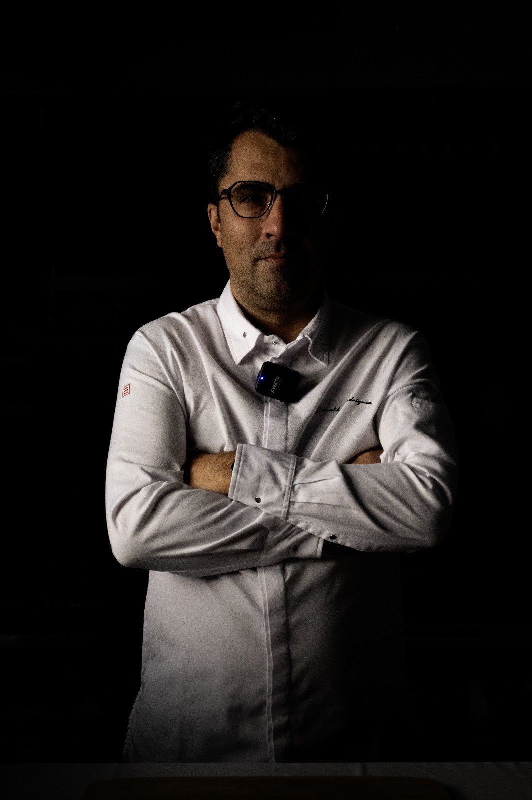 Hombre con gafas y camisa blanca de pie con brazos cruzados en un entorno oscuro.