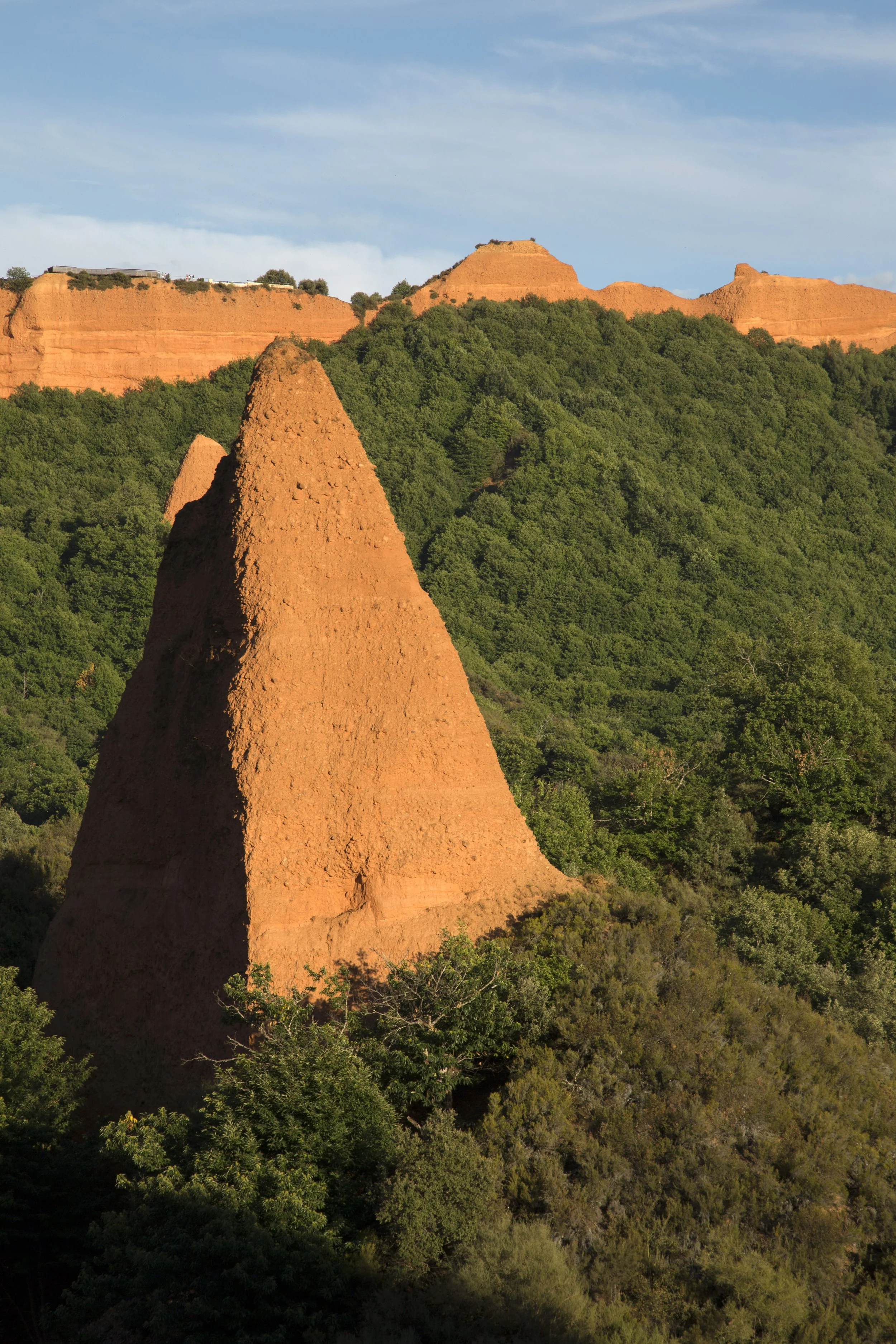 Paisaje de colinas con formaciones rocosas de color terracota y abundante vegetación verde bajo un cielo parcialmente nublado.