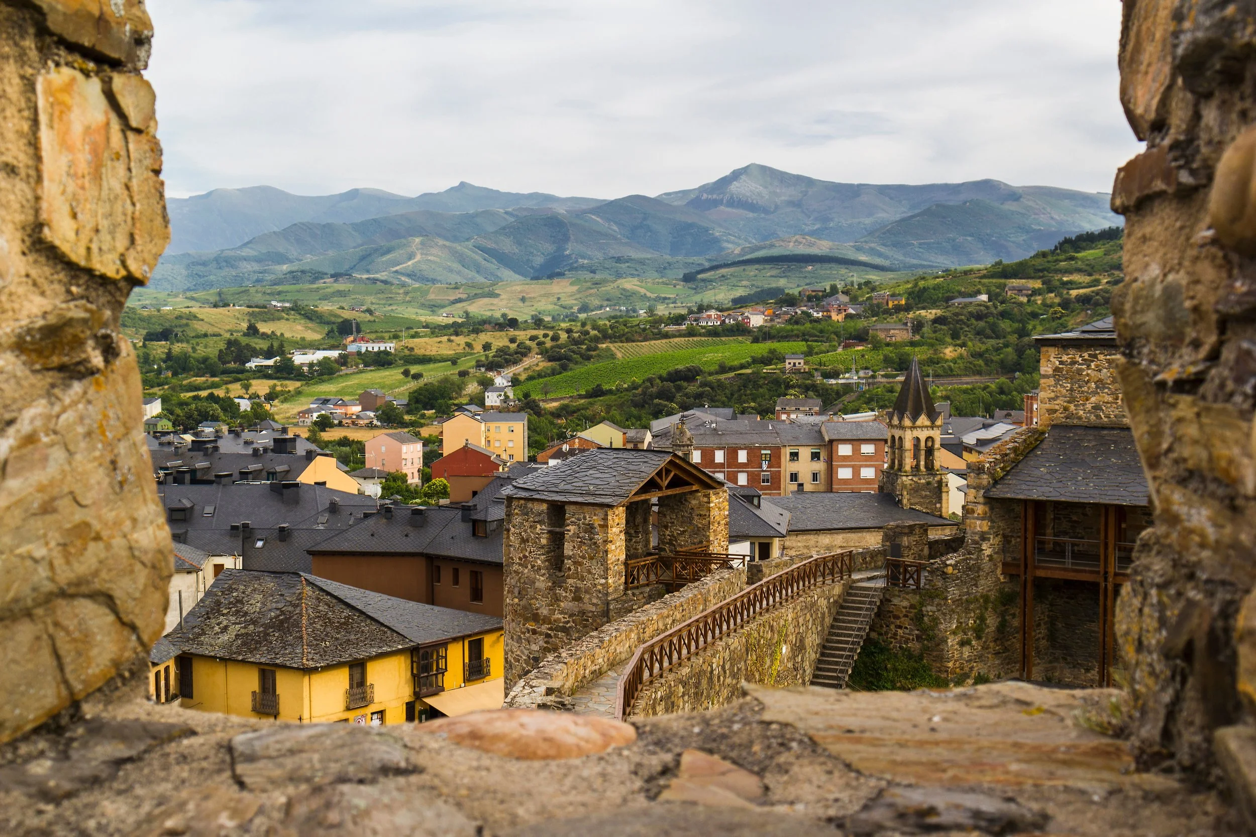 Vista panorámica de un pueblo con casas coloridas y una iglesia, rodeado de campos verdes y montañas al fondo, vista desde las murallas de un castillo o fortaleza.