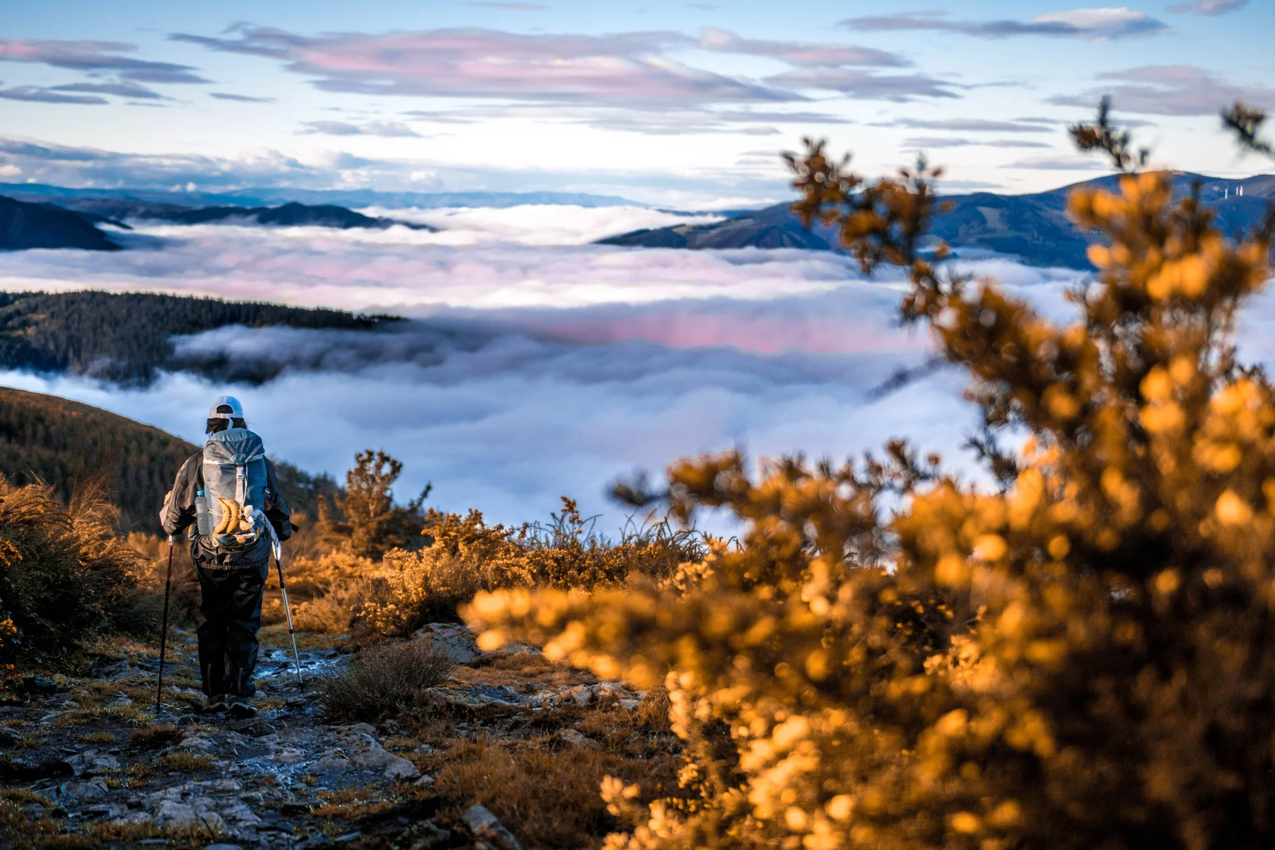 Dos senderistas caminando por un sendero en una cima con vista a una nube en forma de manto sobre las montañas y un cielo con nubes rosas y azules.