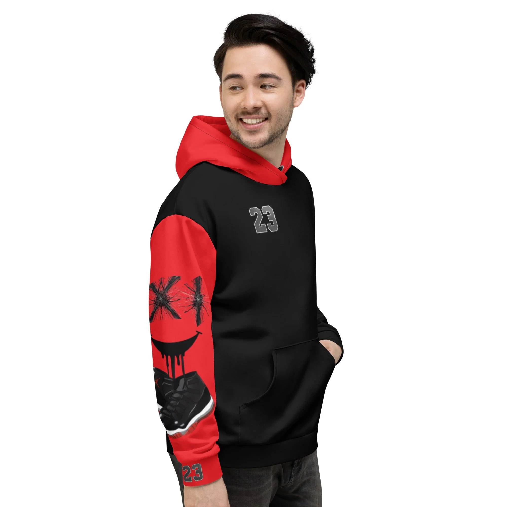 A young man with dark hair and a light beard is wearing a black and red hoodie with a graffiti-style smiley face on the sleeve and the number 23 on the chest and sleeve. He is smiling and looking to his left, with his hands in his hoodie pocket.