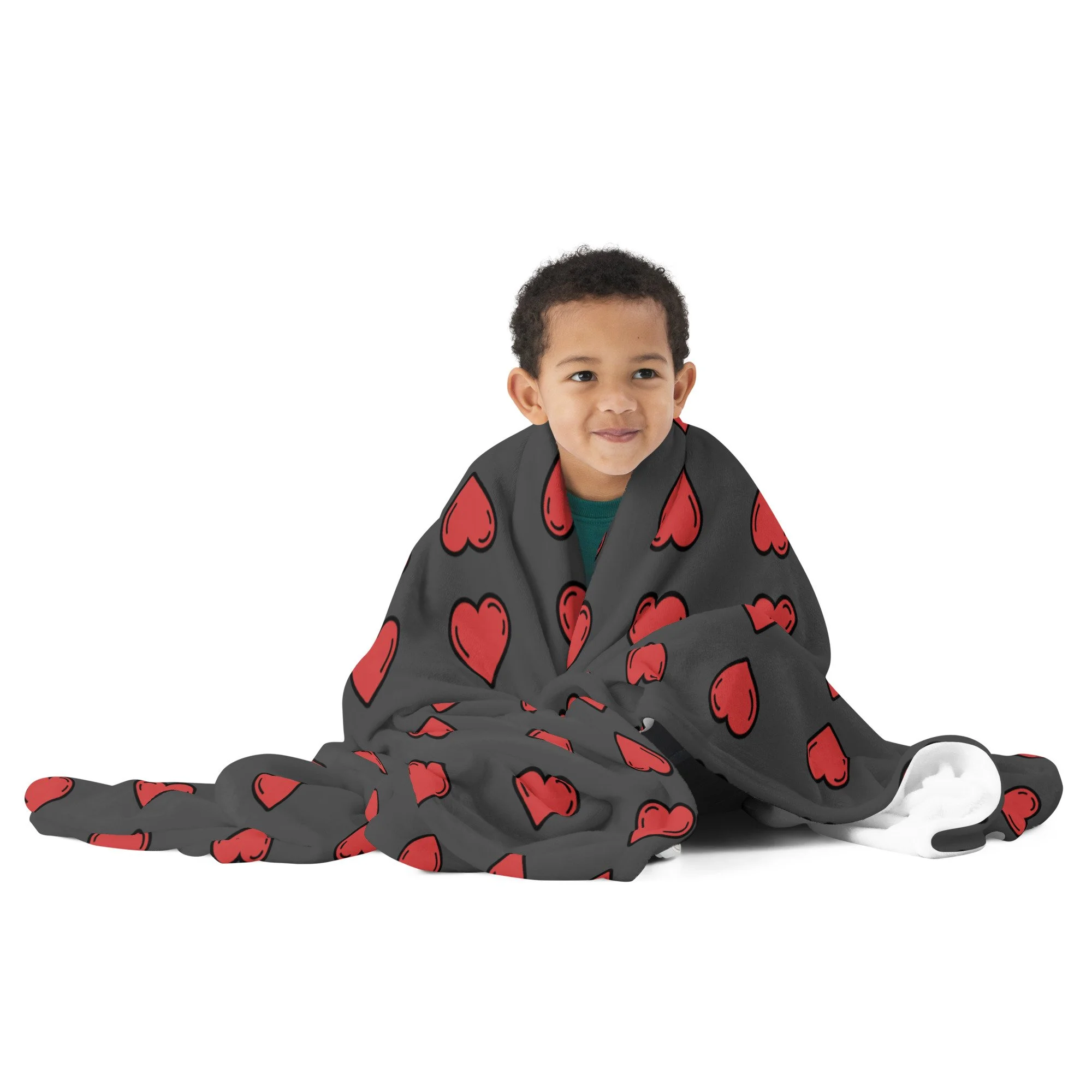 Young boy sitting on the floor wrapped in a blanket with red hearts, smiling, with a white background.