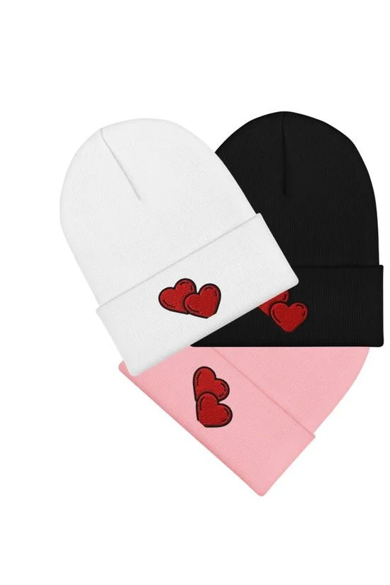 Three knit beanies in white, black, and pink, each with red heart embroidery, arranged in a fan shape.
