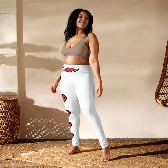 Woman wearing beige crop top and white leggings with hearts, standing indoors on a woven rug, with shadows cast on the wall behind her.
