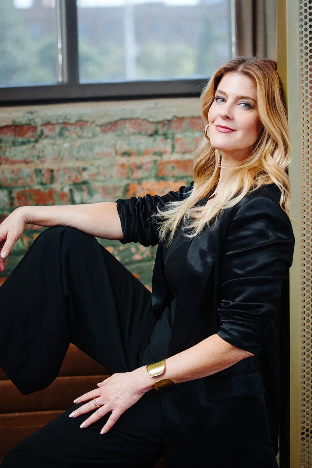 A woman with long wavy blonde hair wearing a black blazer and black pants, sitting indoors with a brick wall and window in the background.