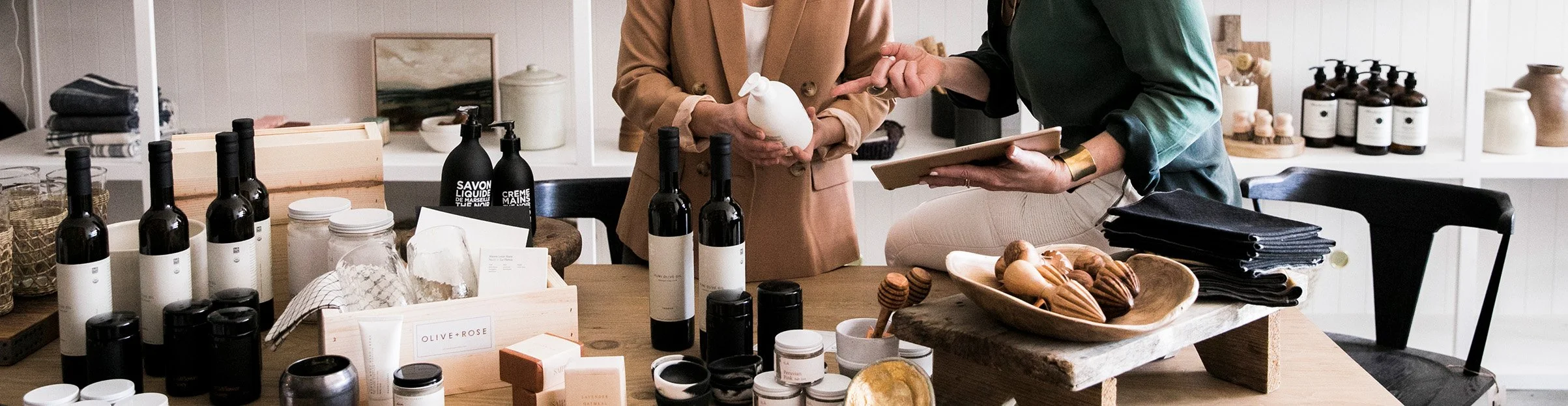 Two women are shopping for personal care products and home goods at a boutique or store, with various bottles, jars, and products displayed on the table and shelves.