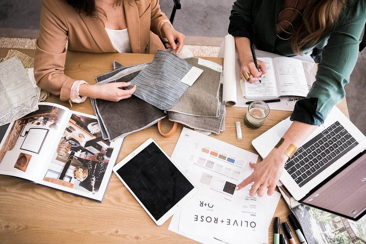 Two women working at a table cluttered with fabric samples, a magazine, a tablet, a notebook, a laptop, and various tools for design or fashion planning.