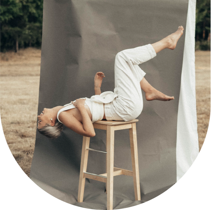 A woman lying on her back on a stool with her legs raised, against a backdrop of gray paper and an outdoor setting with trees and grass.