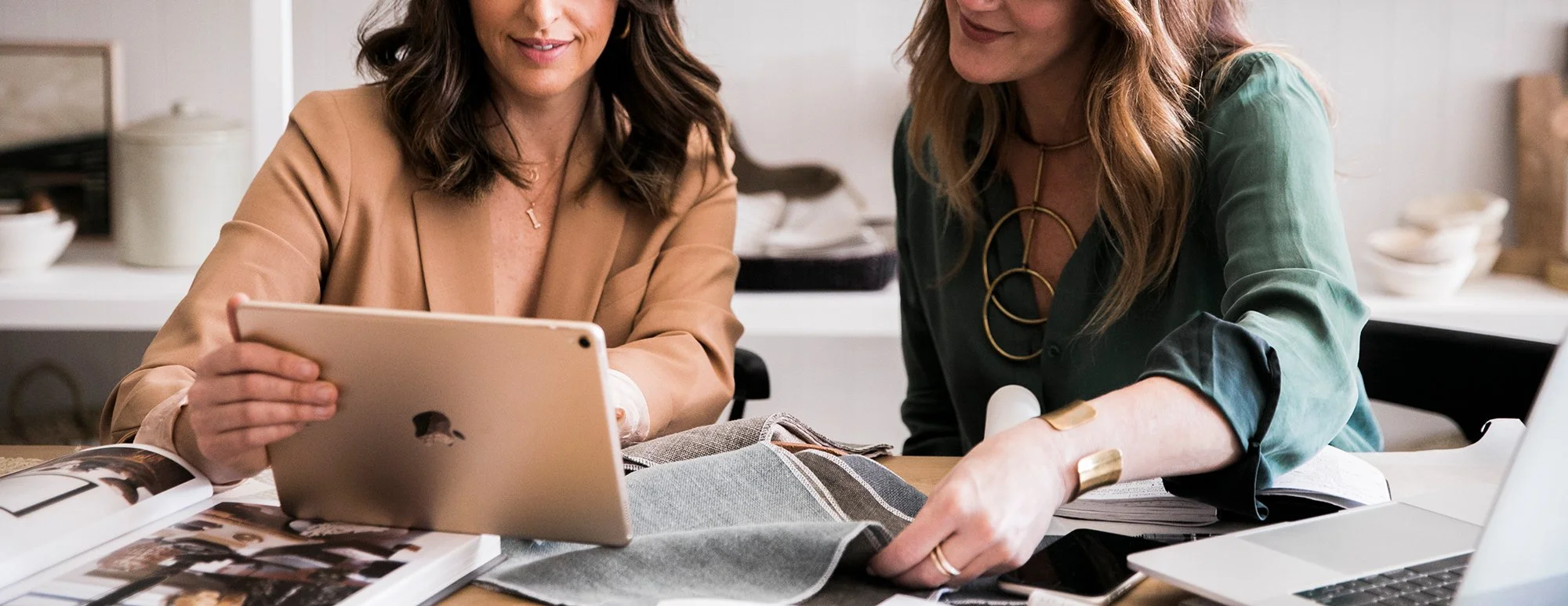 Two women sitting at a table, looking at a tablet together, surrounded by design materials and a laptop.