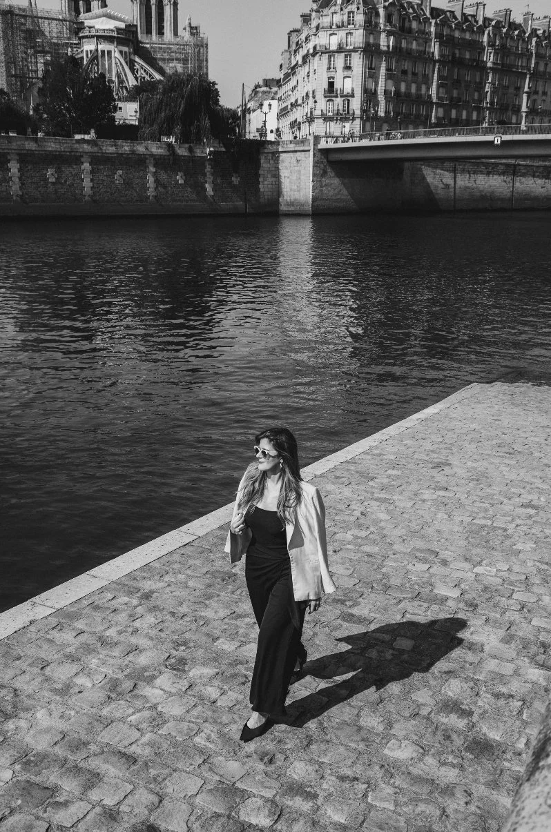 A woman wearing black pants, a black top, a light-colored blazer, and sunglasses walking along a cobblestone pathway by a river, with city buildings and a bridge in the background.