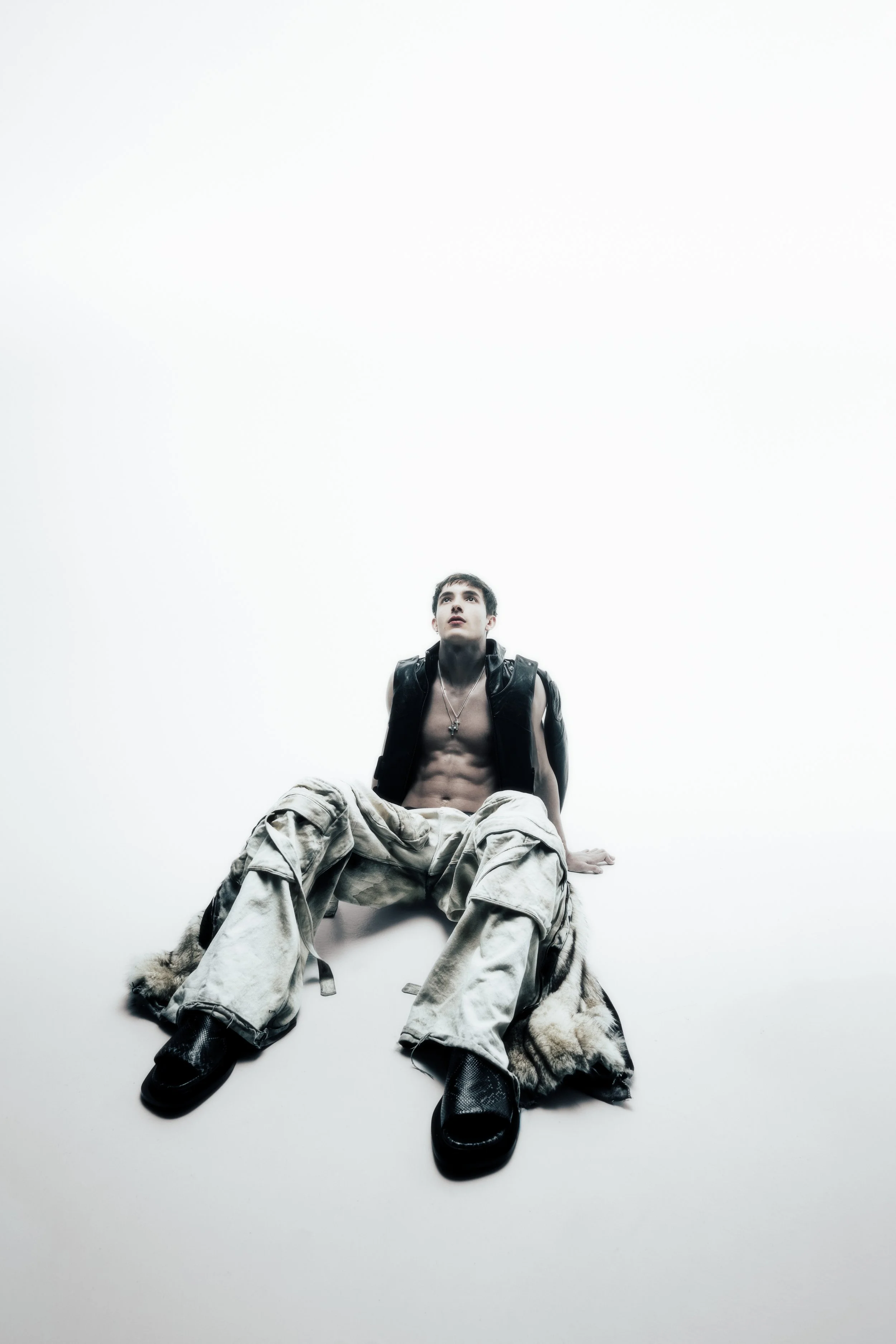 Young man sitting on the floor with his legs spread, shirtless with a black vest, wearing baggy camouflage pants and black shoes, looking upward against a plain white background.