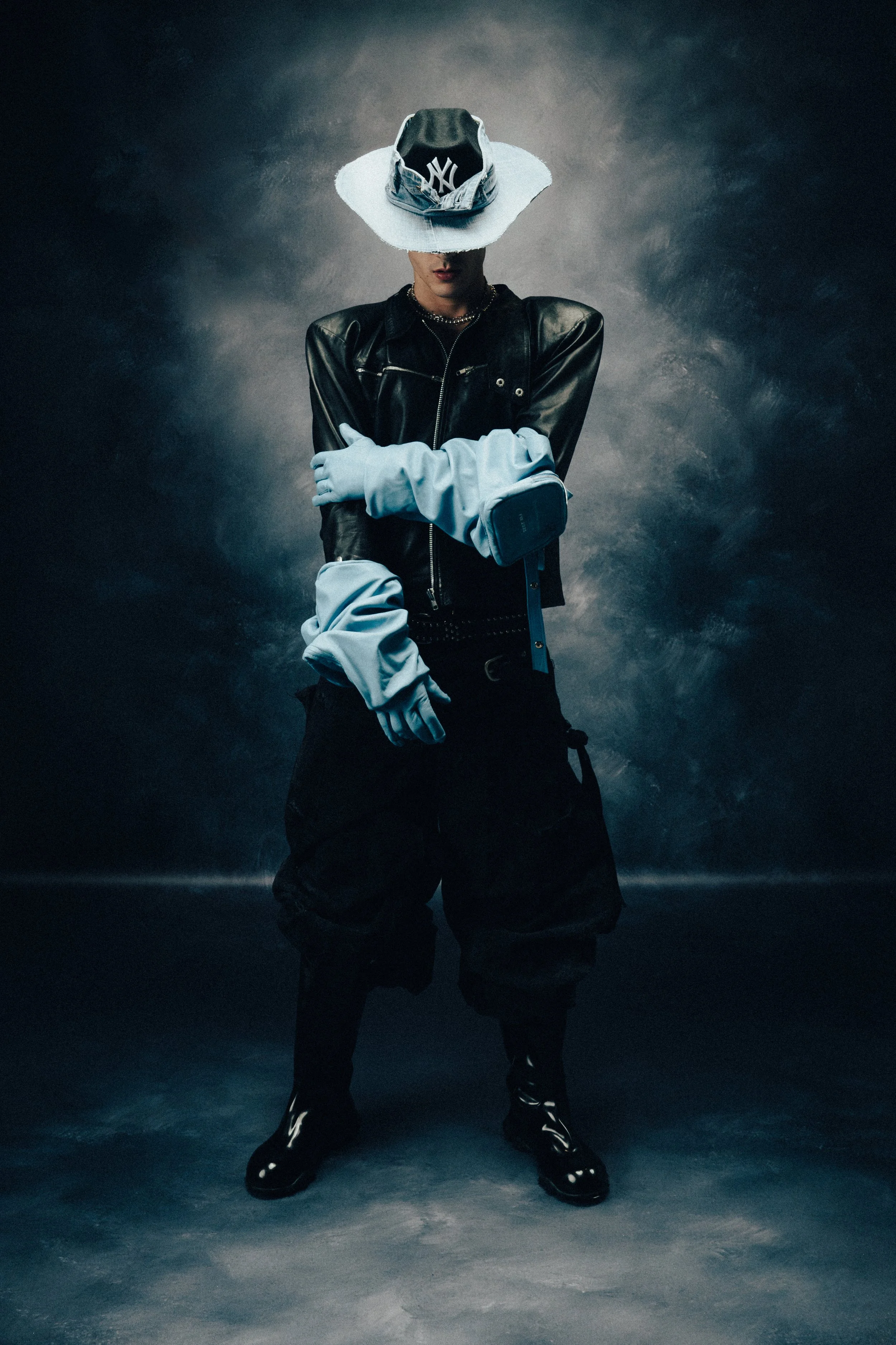 A person dressed in black pants, a black leather jacket, white gloves, and shiny black boots, wearing a large white wide-brimmed hat with a denim pattern and a New York Yankees cap underneath, against a dark textured background.