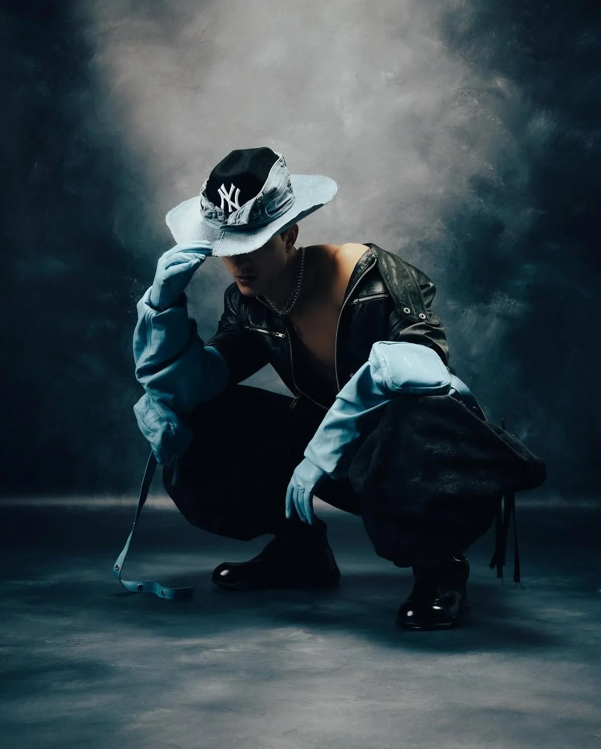 A person crouching, wearing a large hat with a New York Yankees logo, sunglasses, a black leather jacket off the shoulders, blue gloves, dark pants, and shiny black shoes, against a dark, textured background.