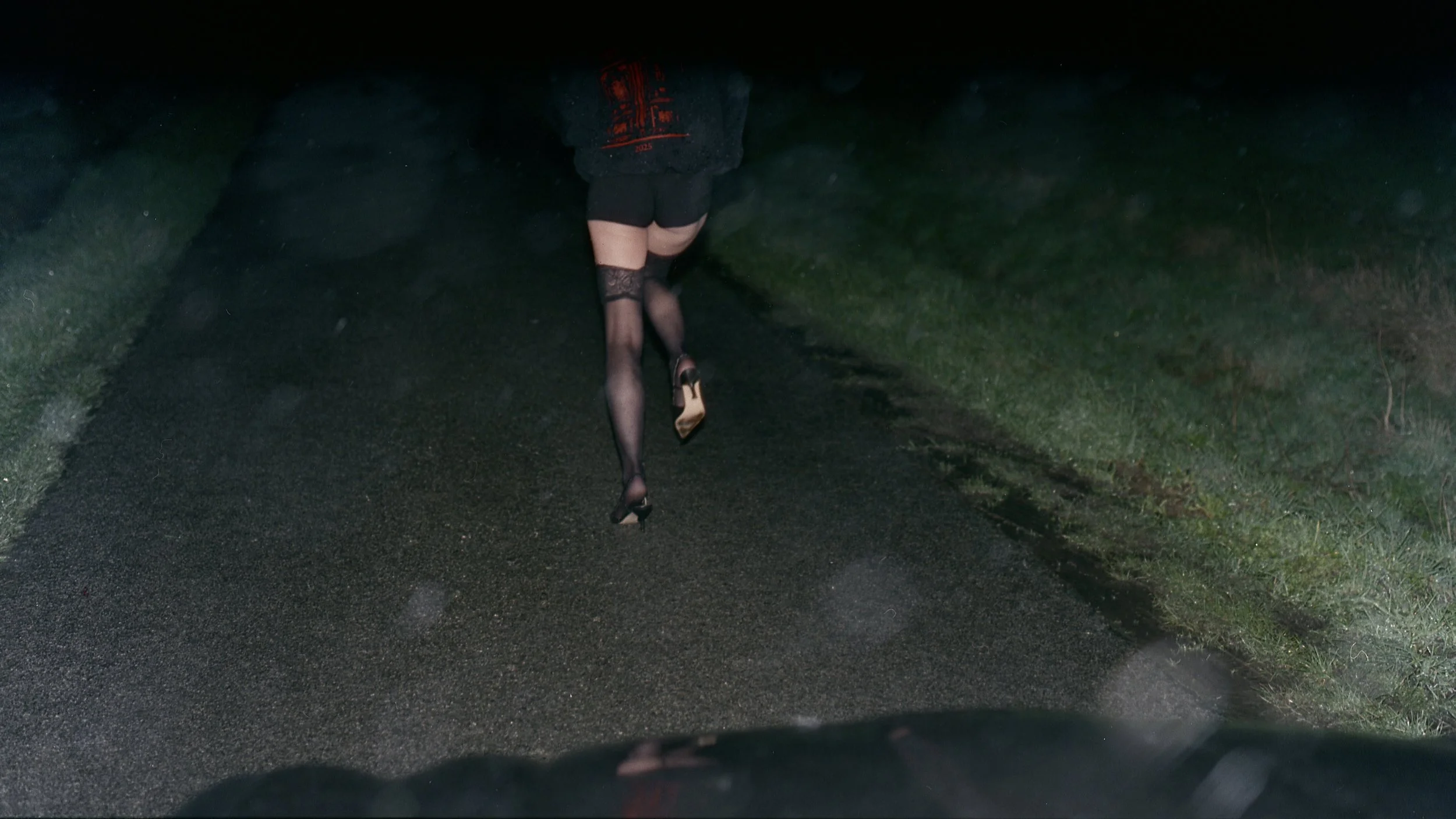 Person wearing stockings and high heels walking on a dark, wet road at night, with green grassy area on the side.