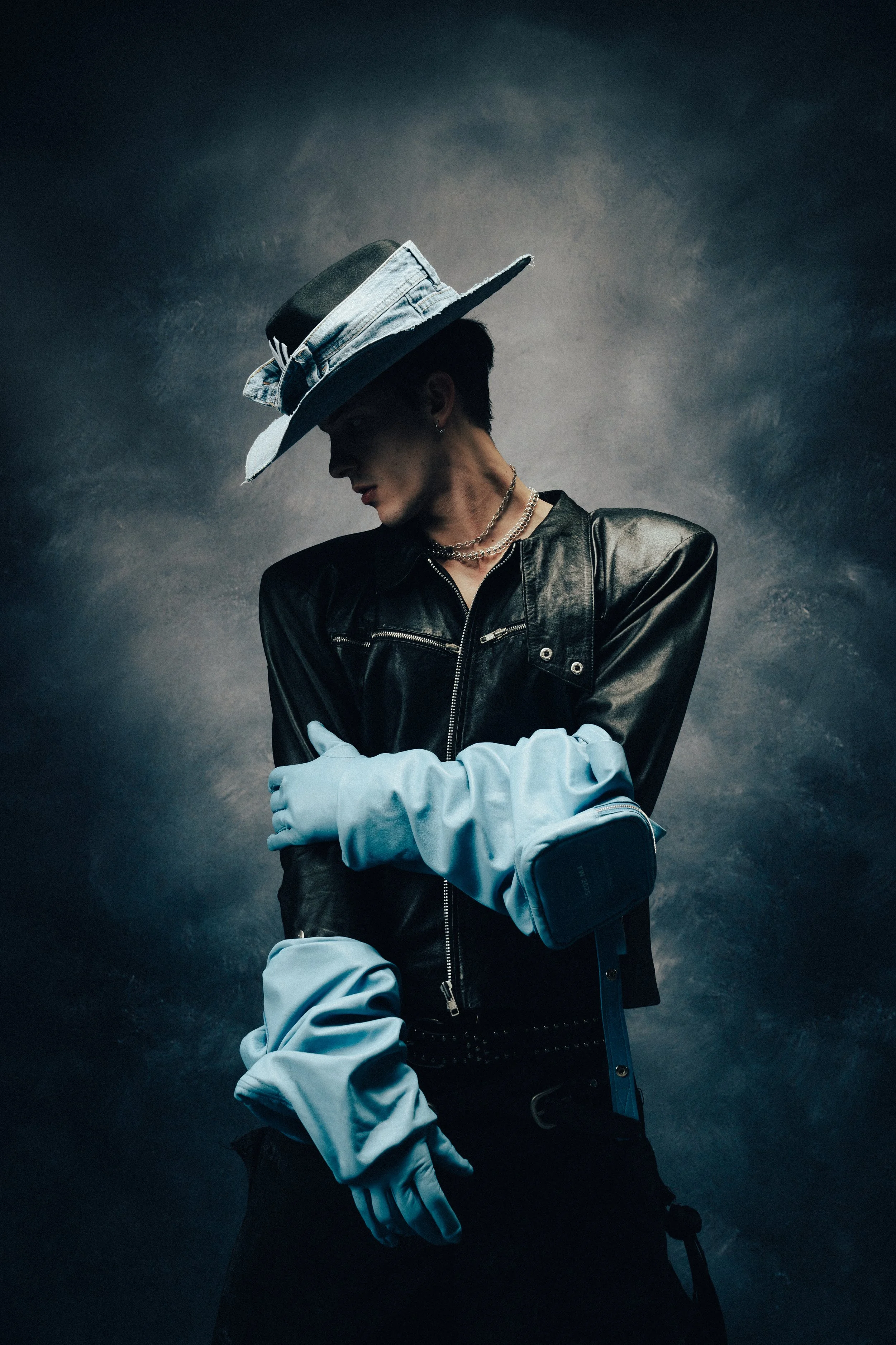A person wearing a black leather jacket, layered necklaces, a hat made from denim with a cigarette in the brim, and light-colored gloves, against a dark textured background.