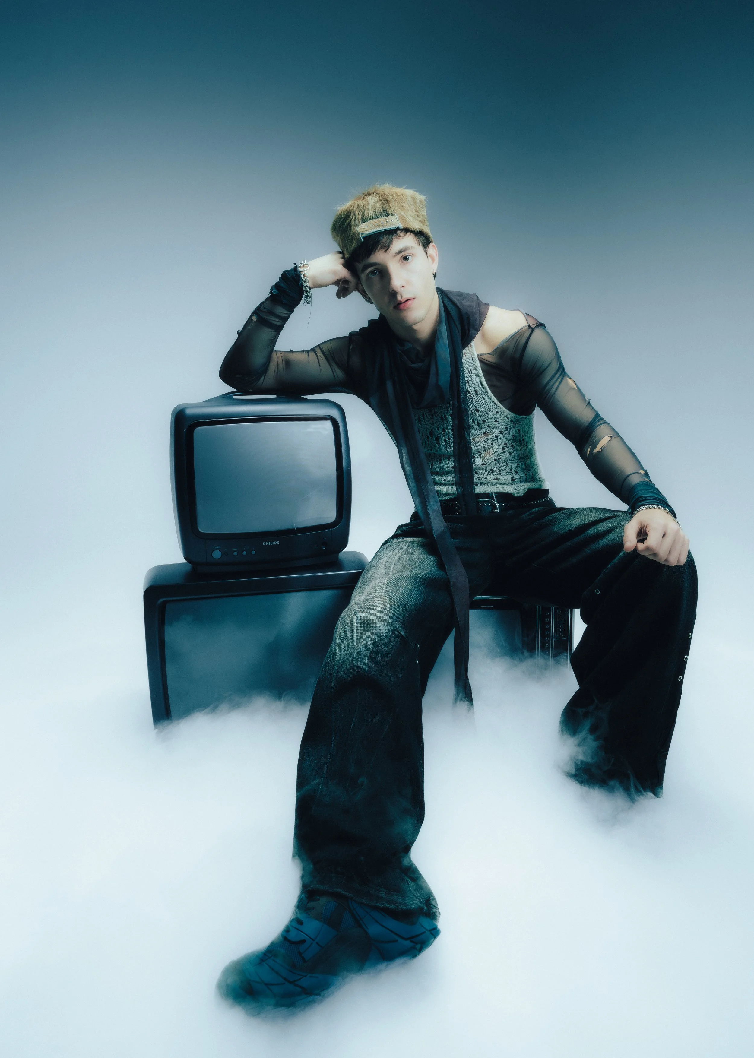 A young person with short, styled hair sitting on a monitor in front of a vintage CRT TV, with fog or smoke on the floor. They are dressed in a sheer, long-sleeve top with a metallic graphic underneath, black baggy pants, and sneakers. They are resting their head on one arm, looking at the camera with a neutral expression.