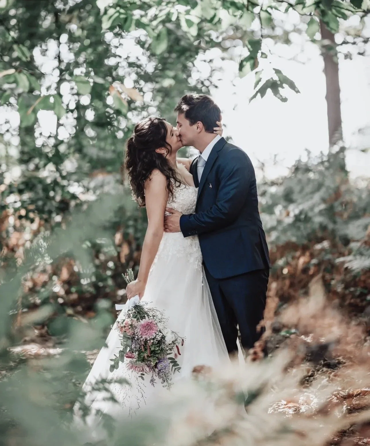 A bride and groom kiss in a wooded outdoor setting, with the bride holding a bouquet of flowers in one hand and the groom in a suit.