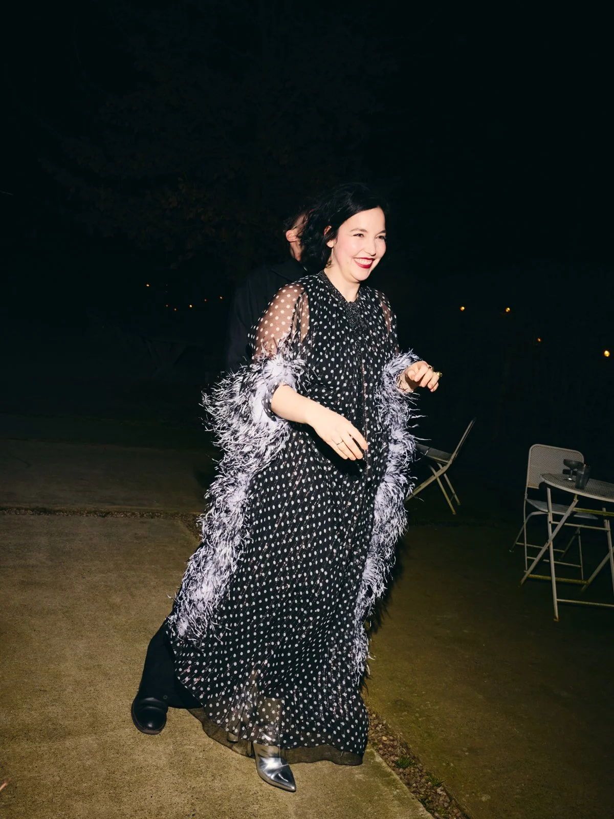 A woman wearing a black polka dot cocktail dress and a feather-trimmed coat, smiling and walking outdoors at night.