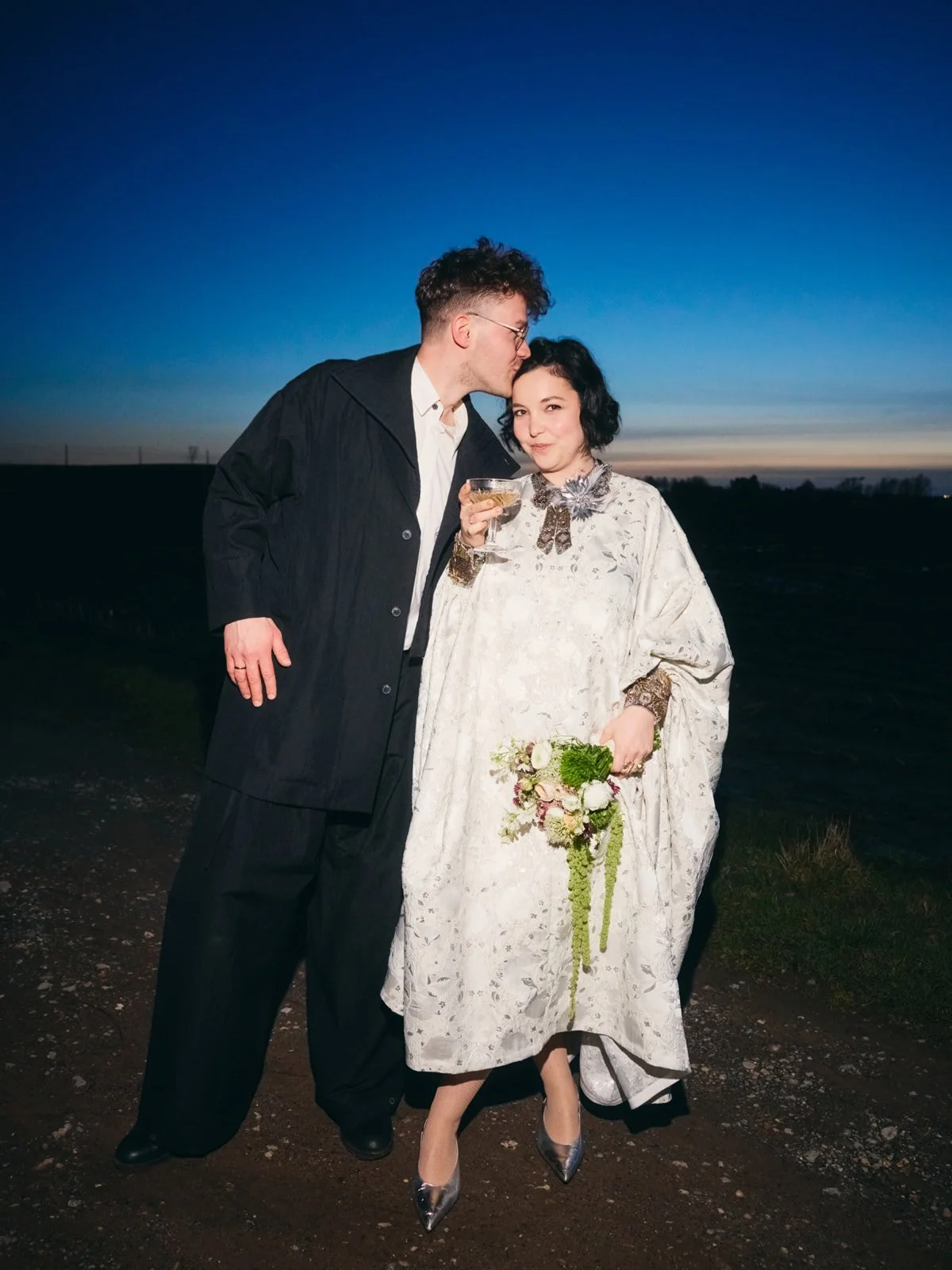 A man and woman standing outdoors at dusk, with the man kissing the woman's forehead. The woman is holding a glass and a bouquet of flowers, dressed in a vintage-style white dress with metallic high heels. The man is wearing a black coat over a white shirt and black pants.